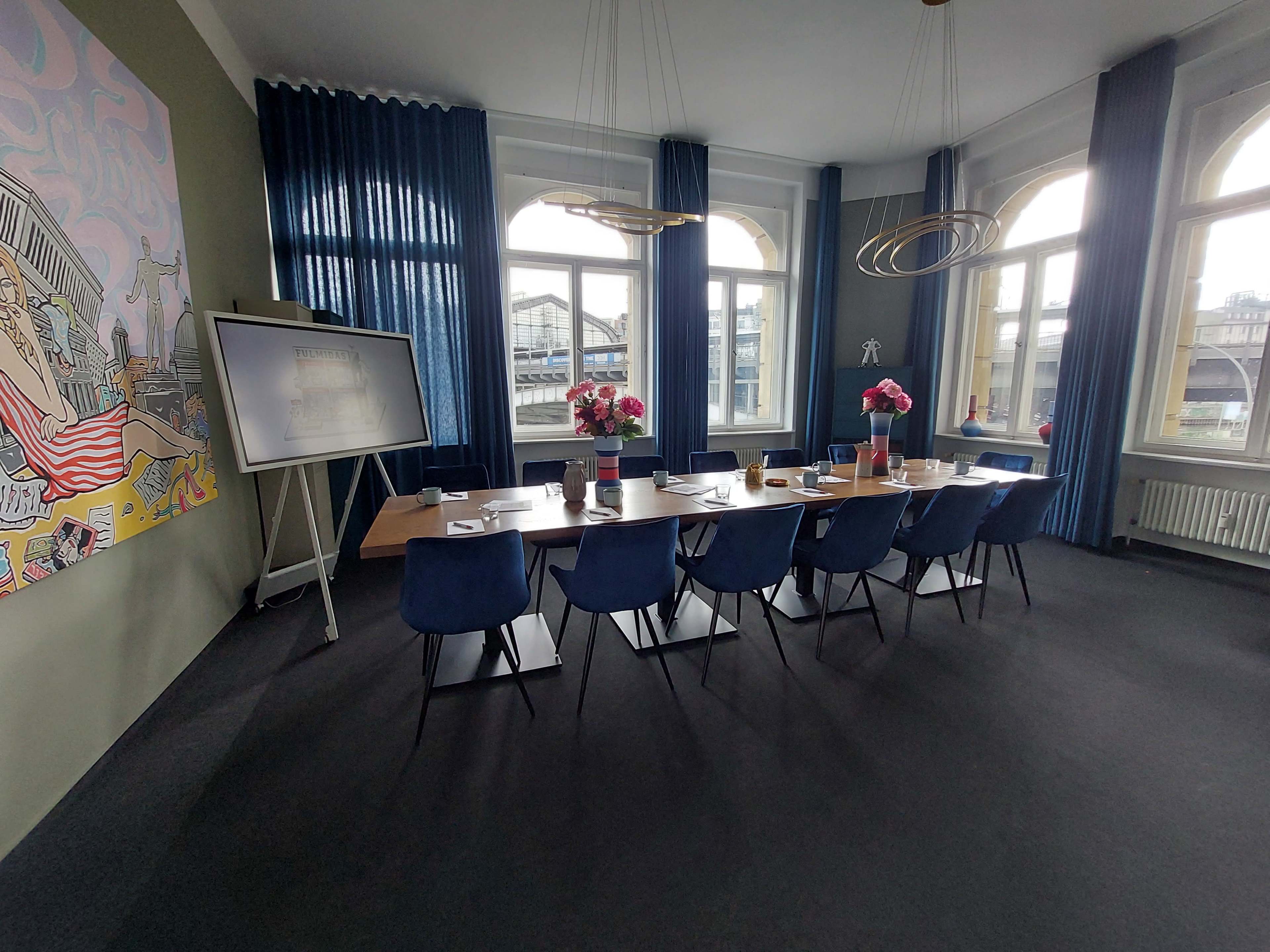 A modern conference room with a long table set for a meeting, surrounded by blue chairs and large windows letting in natural light.