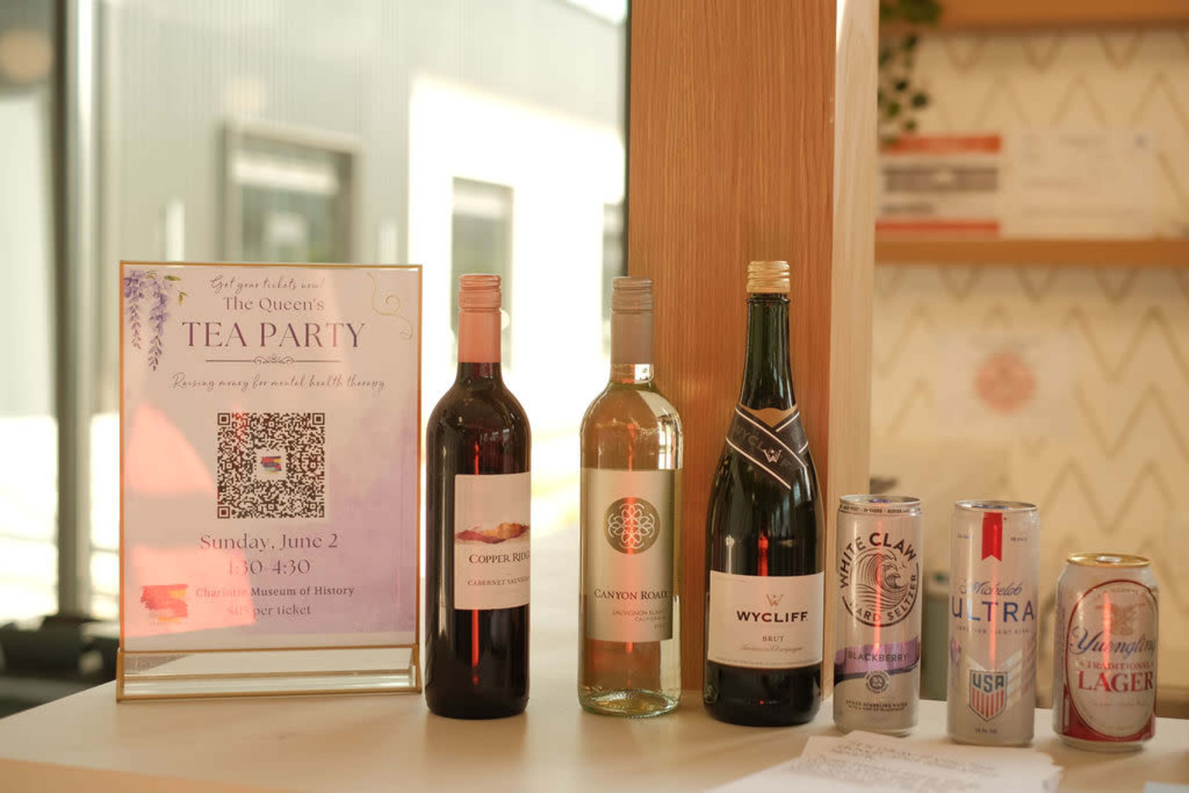 The image displays a table with a sign for "The Queen's Tea Party" alongside bottles of red and white wine, as well as cans of various beers.