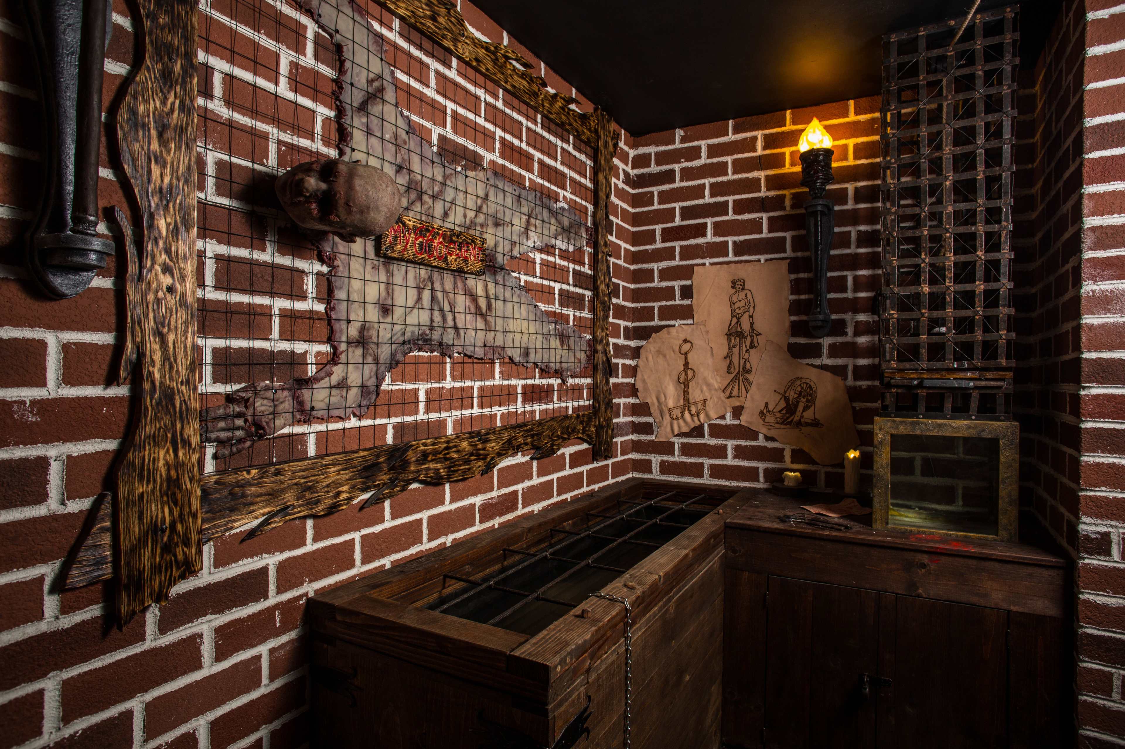 A dimly lit room with brick walls, featuring a macabre display of a skull on a wooden frame, along with eerie drawings and a wooden chest in the corner.
