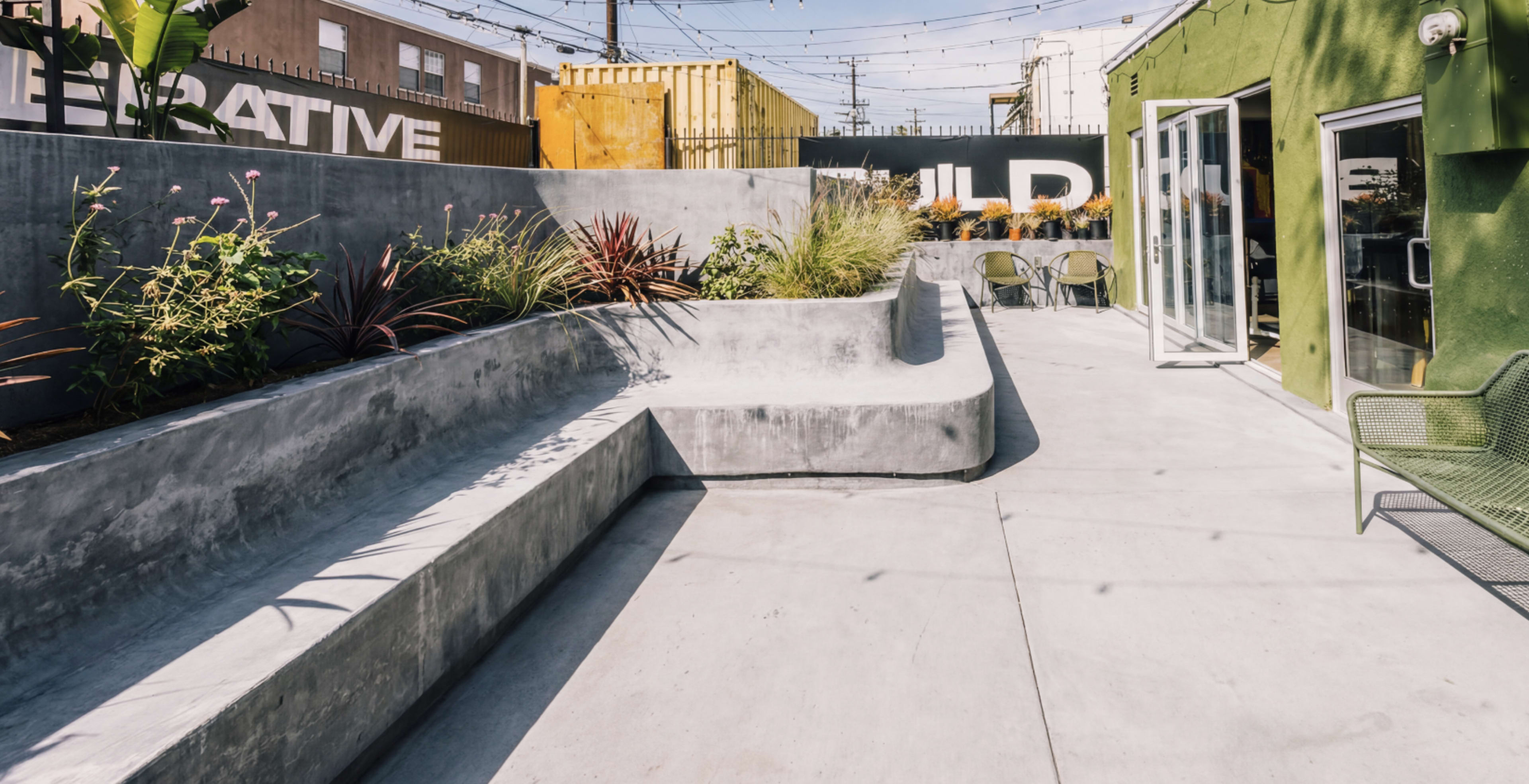 The image shows a concrete outdoor space with landscaped planters and seating areas, surrounded by buildings.