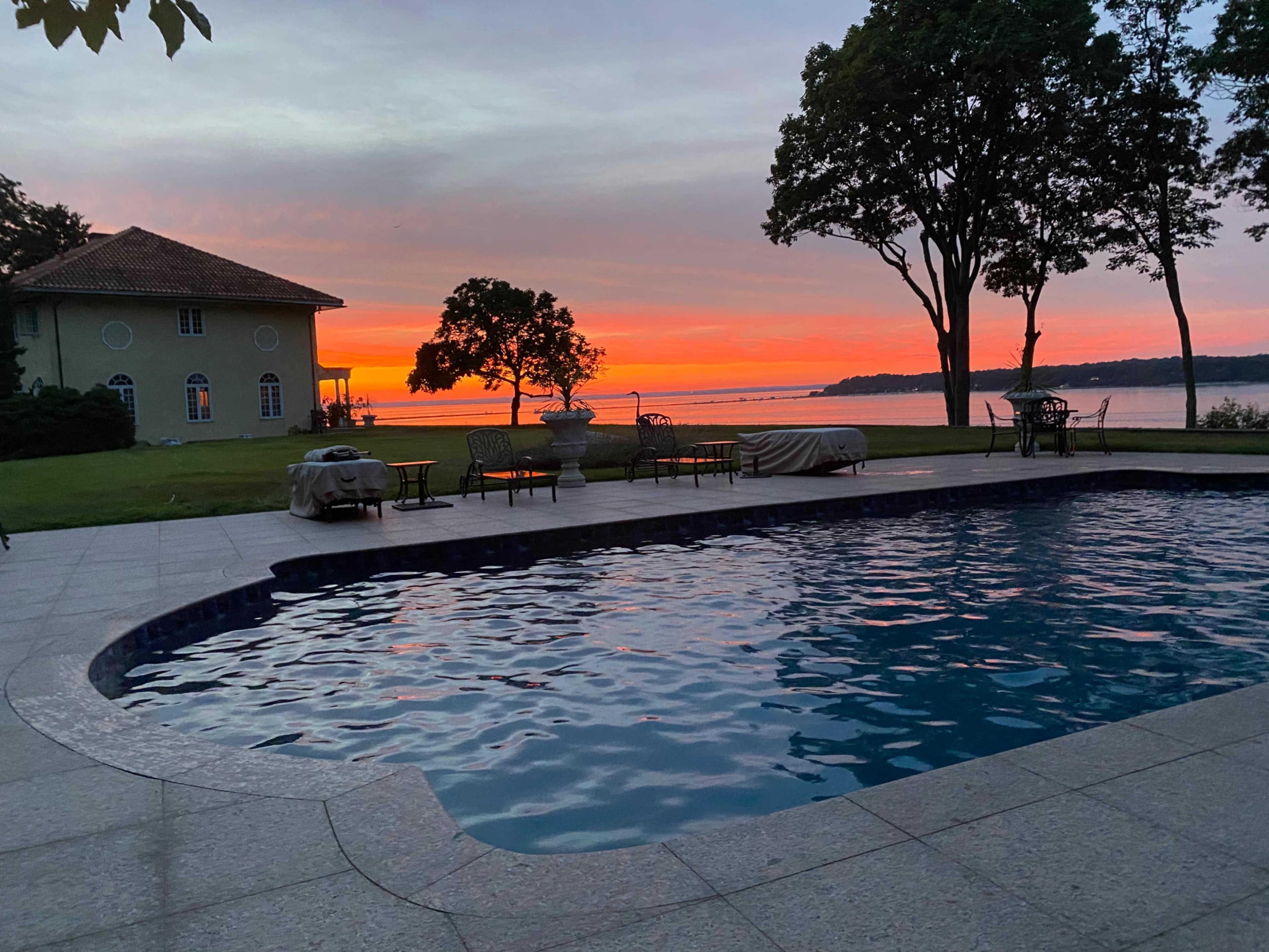 A swimming pool sits in the foreground with a view of a sunset sky over a body of water, framed by trees and a house in the background.