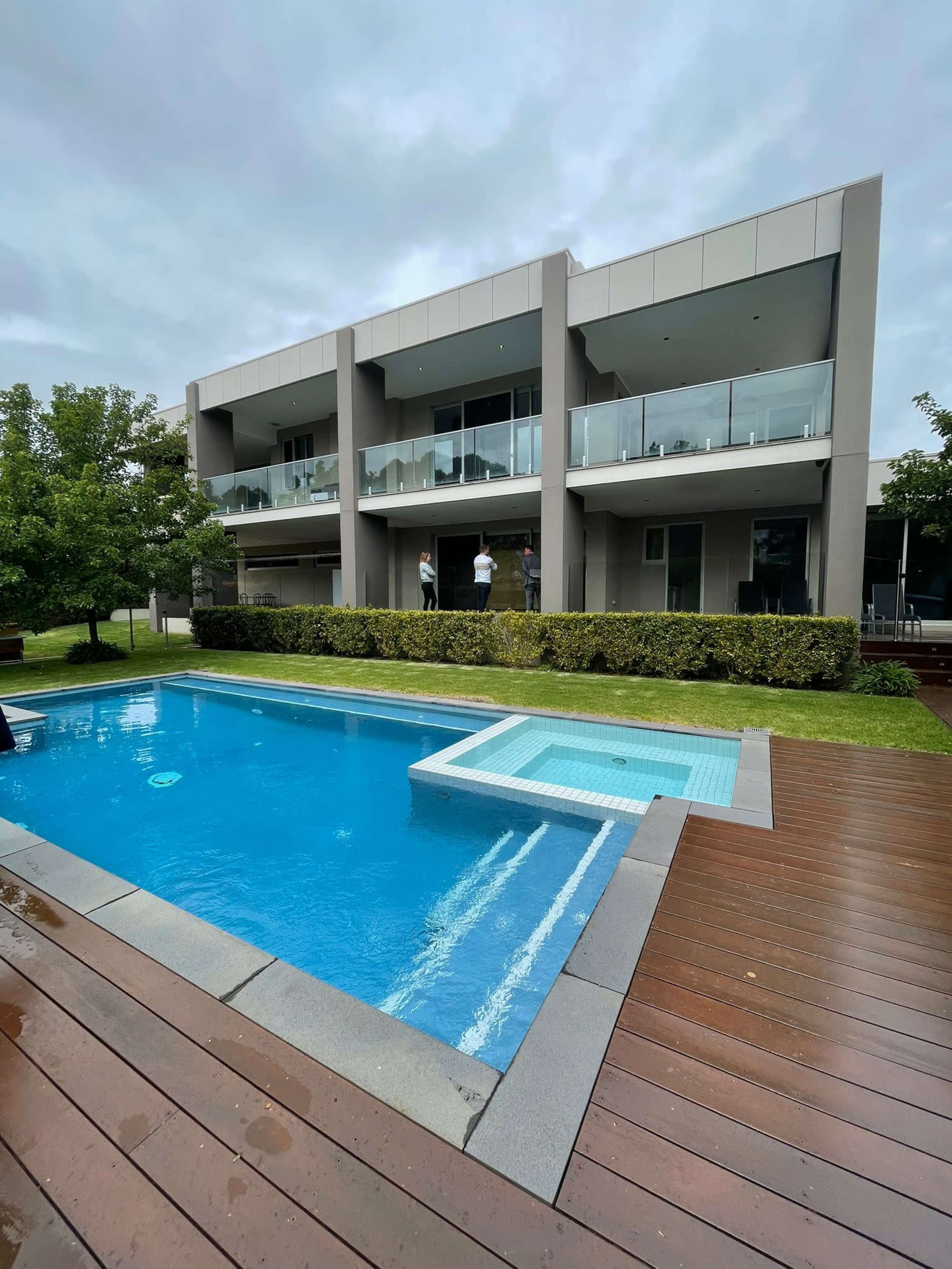 A modern two-story building with balconies overlooks a swimming pool and spa surrounded by grass and wooden decking.