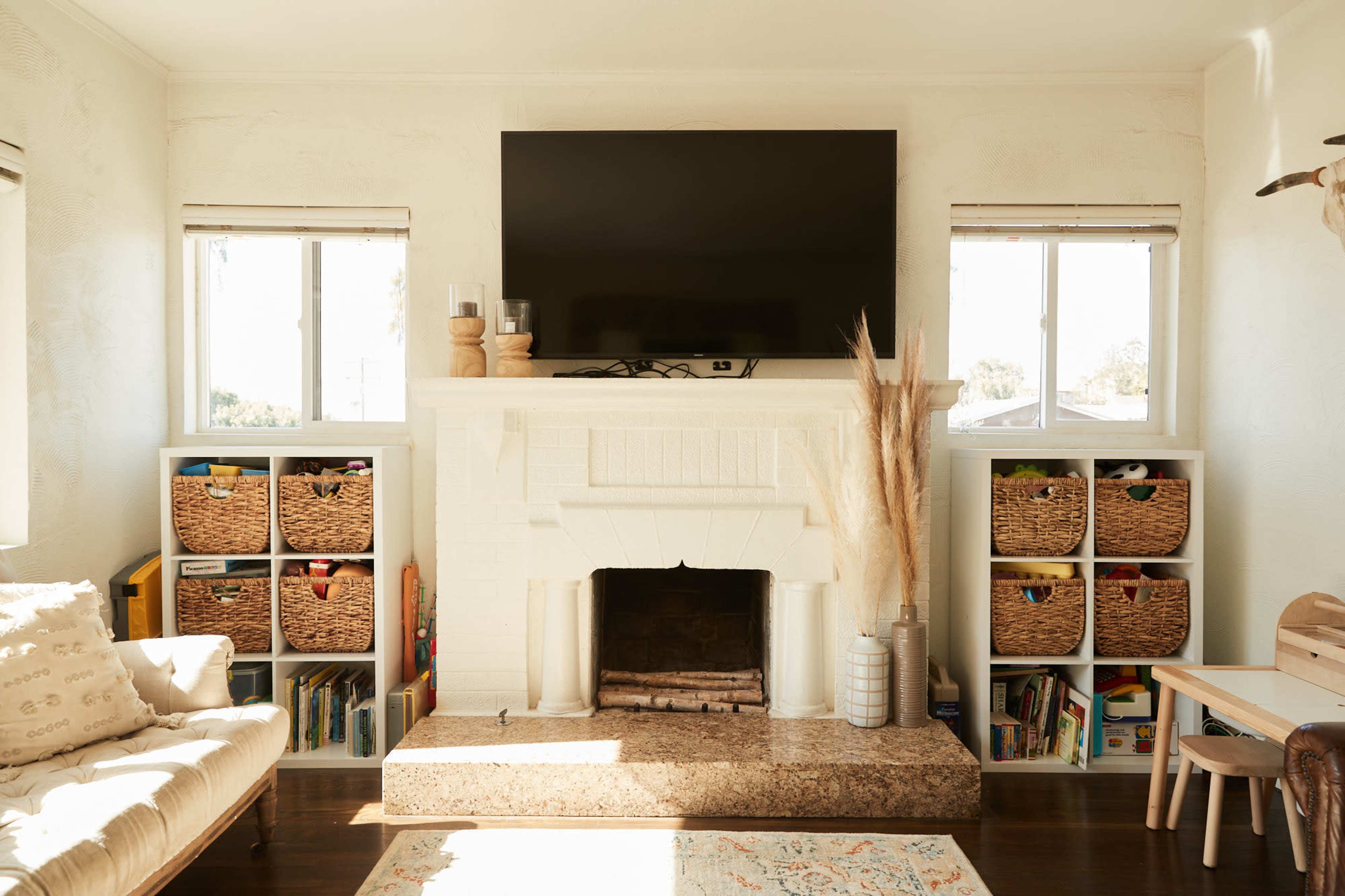 The living room features a white-brick fireplace beneath a wall-mounted TV, surrounded by shelving units filled with baskets and children's books.