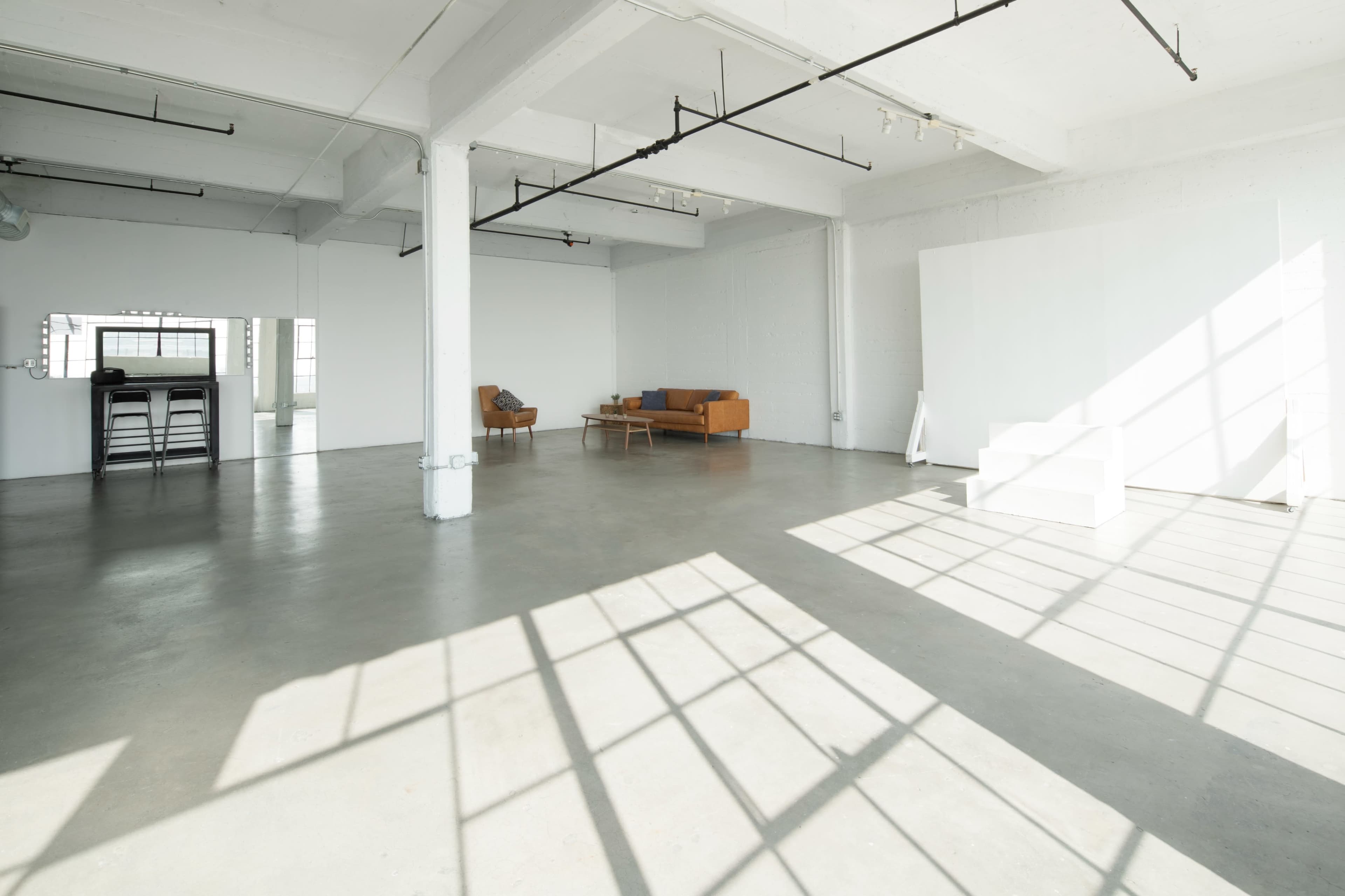The image shows a spacious, minimalist studio with concrete floors, large windows, and a few pieces of furniture including an orange couch and a coffee table.