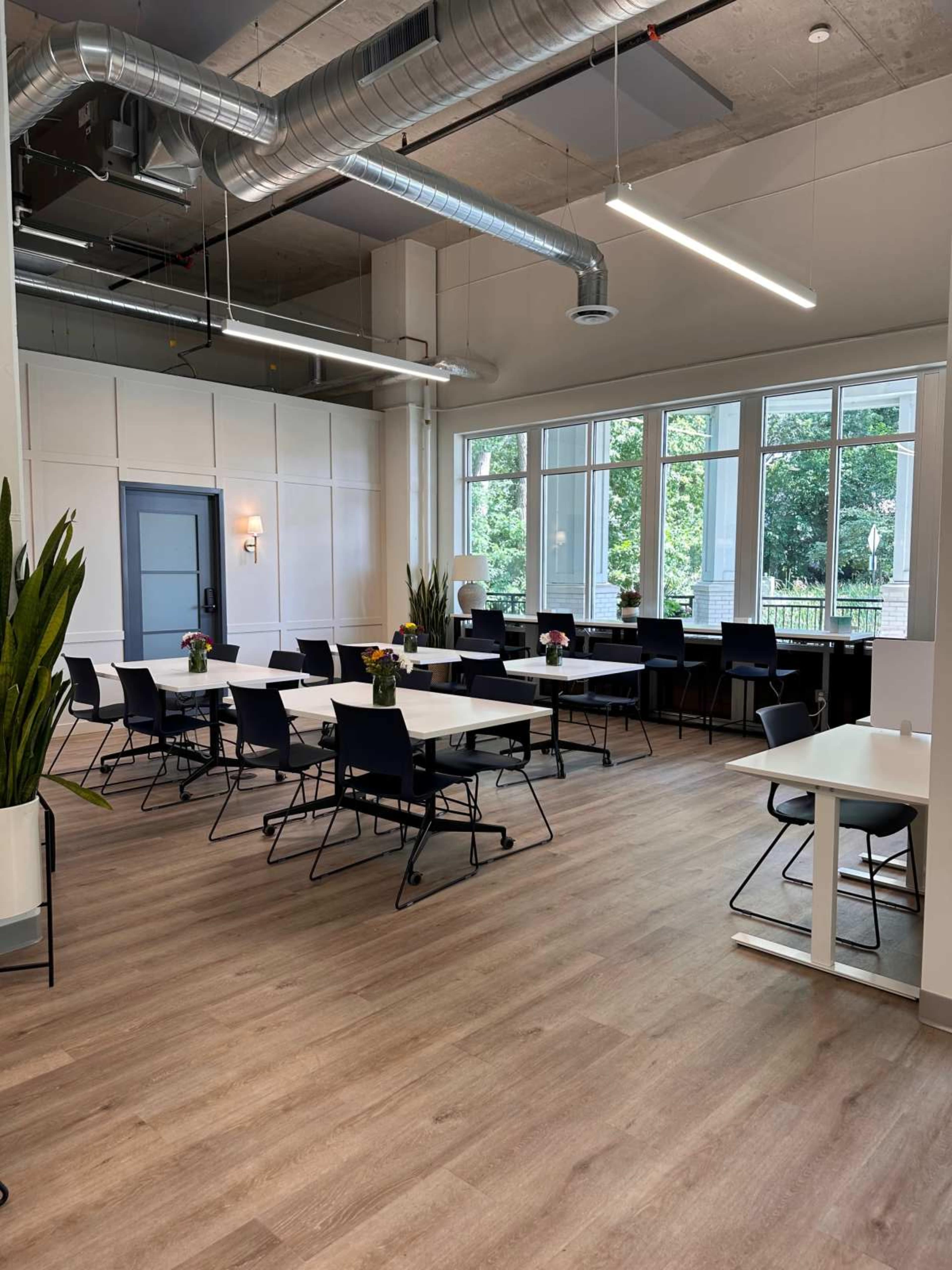 The image shows a spacious, modern room with rows of tables and chairs, large windows providing natural light, and potted plants strategically placed around the space.