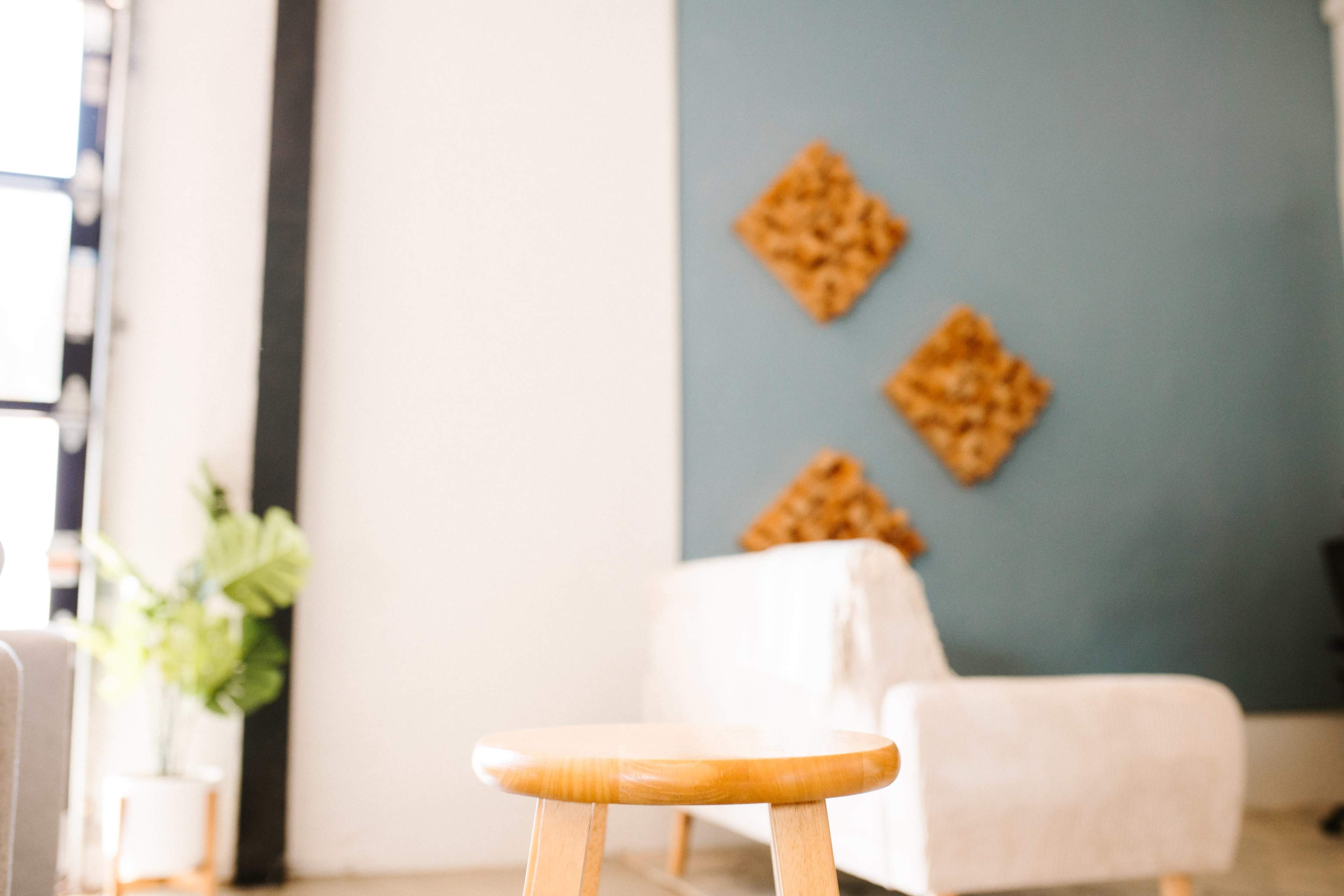 A wooden stool is positioned in the foreground of a bright room featuring a sofa and textured wall art.