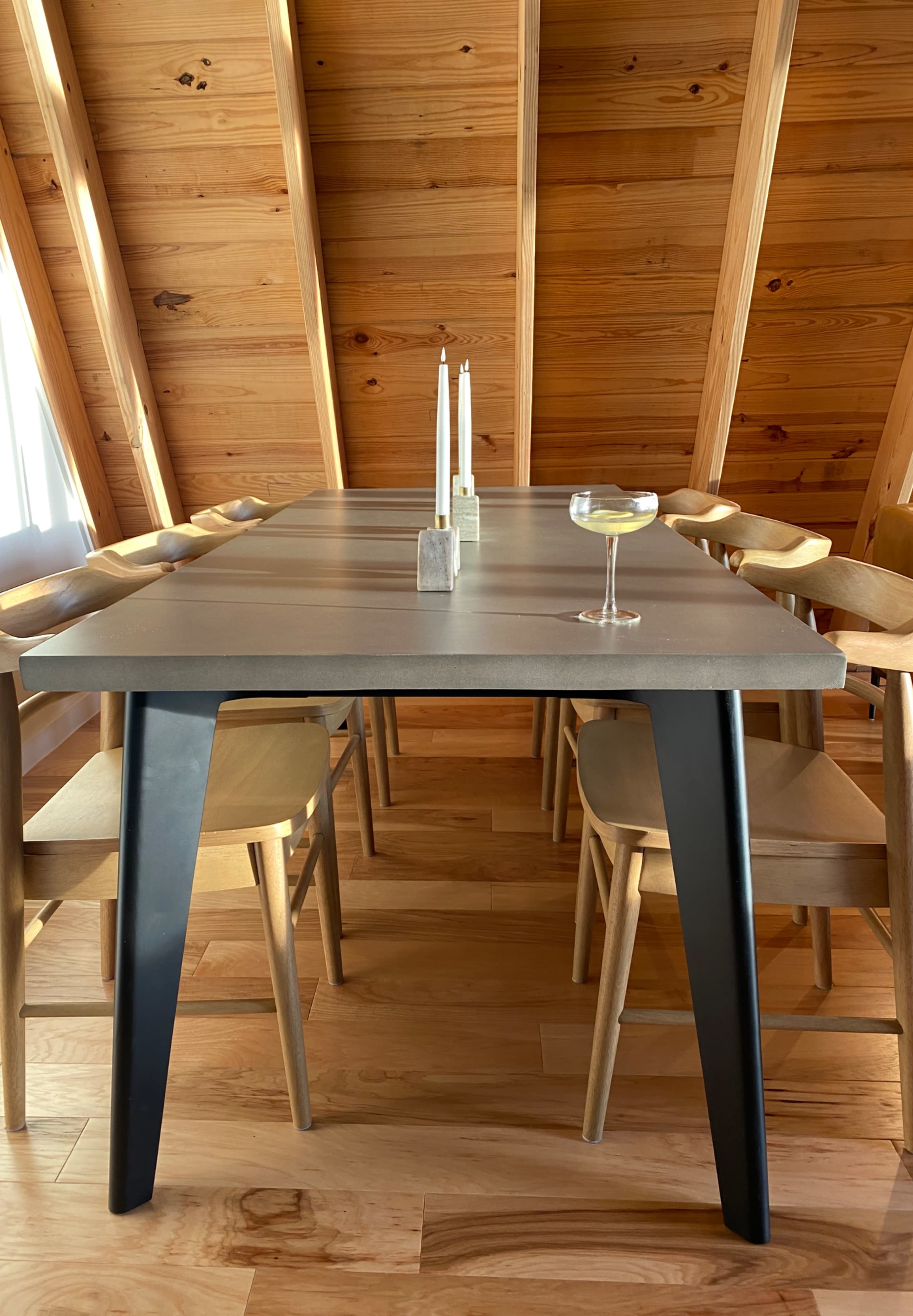 A modern dining table with a gray surface and black legs is set with a cocktail glass and flanked by wooden chairs in a room with wooden walls and ceiling.