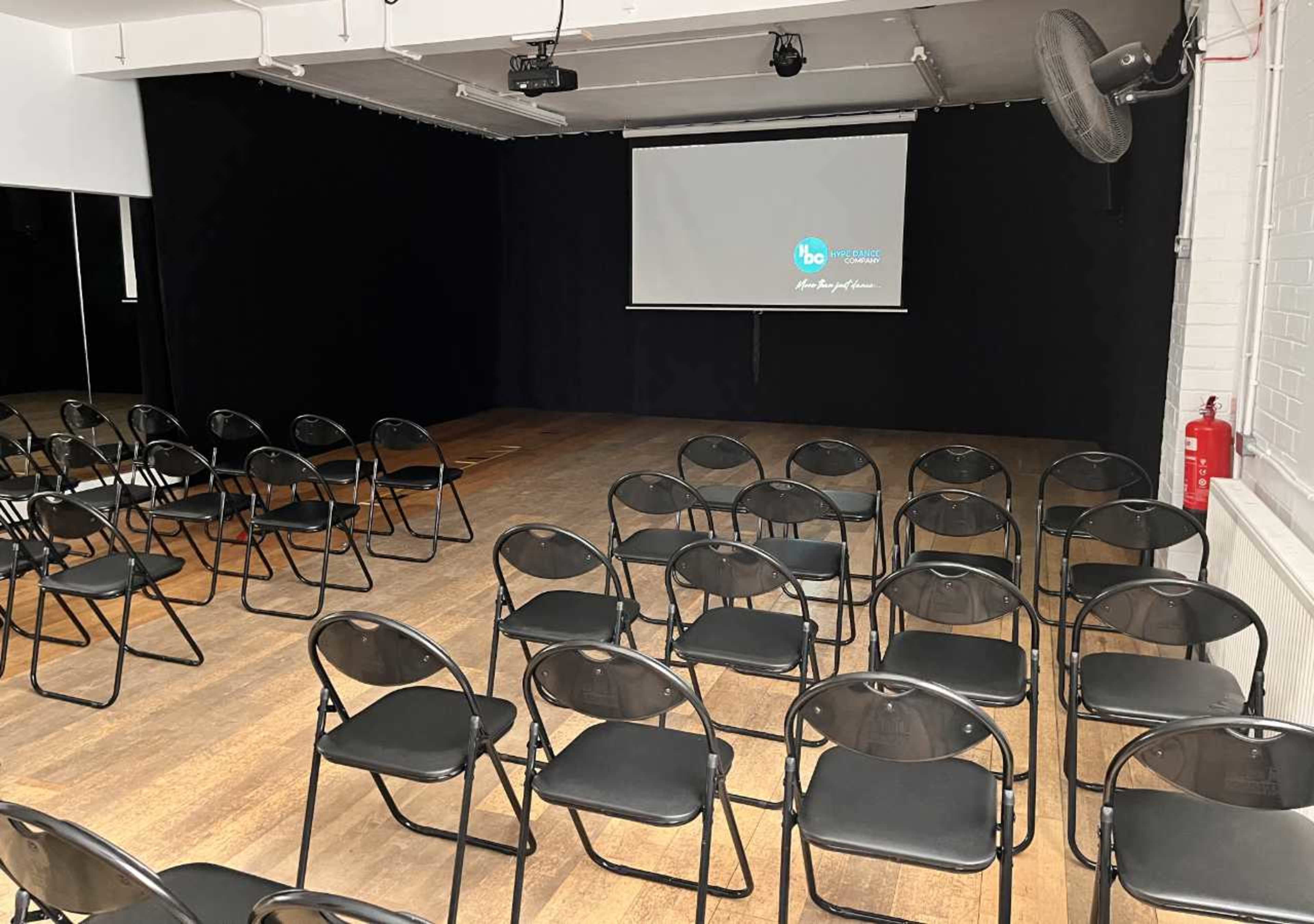 The image shows a spacious room set up with rows of black chairs facing a large screen against a dark backdrop.