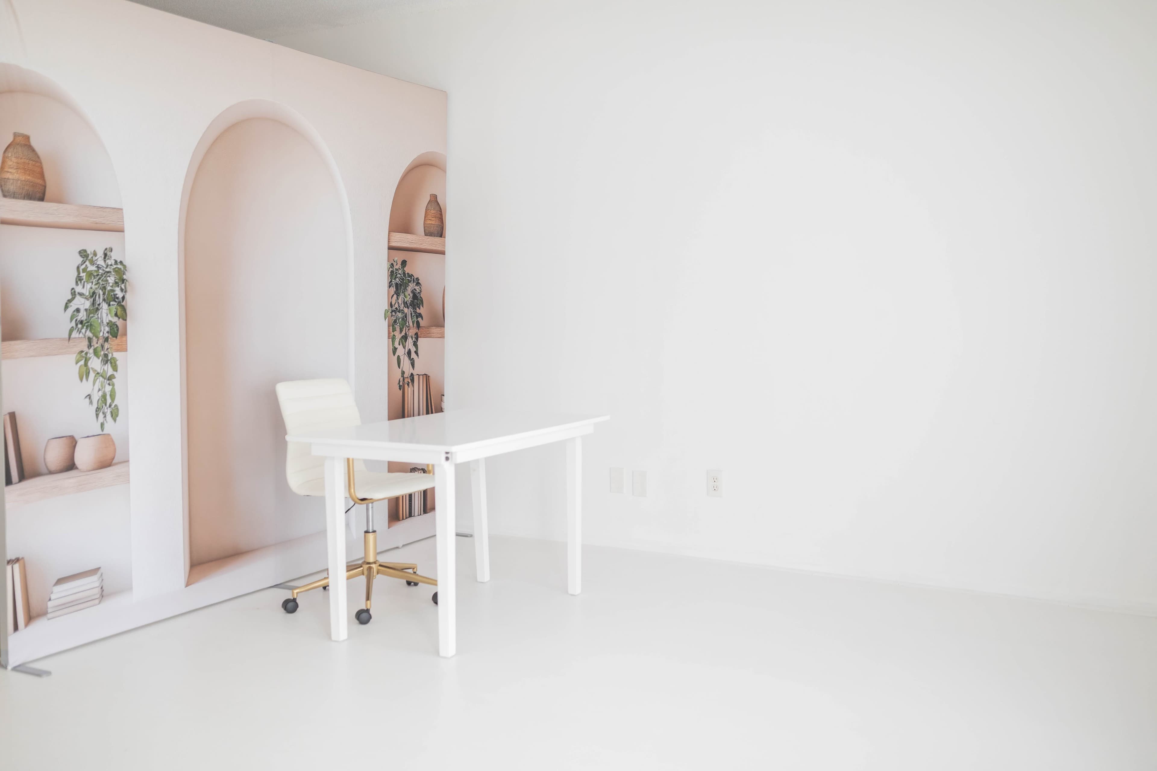 The image shows a minimalistic office space featuring a white desk with a rolling chair against a pale background with arch-shaped shelves.