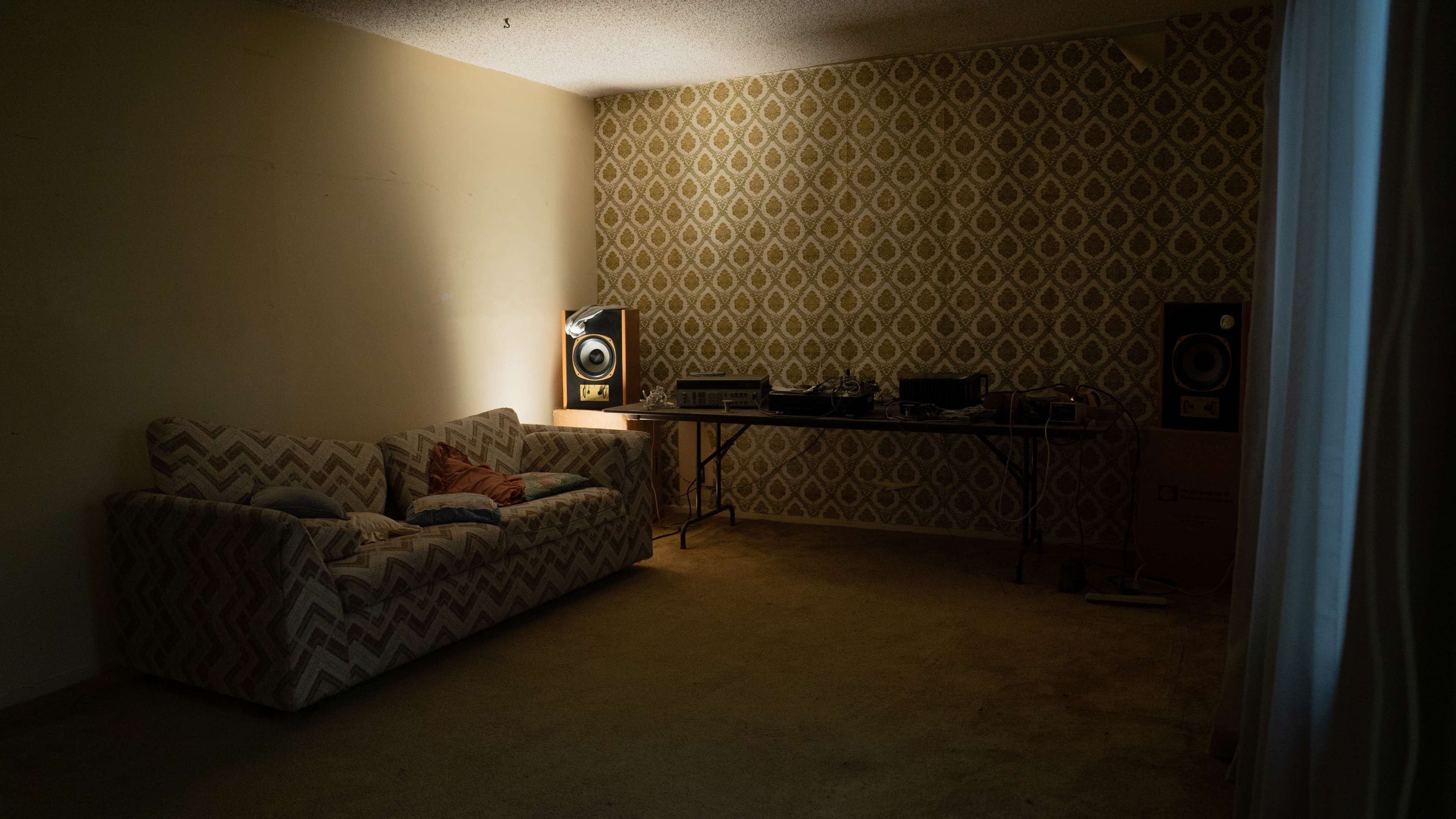 The image shows a dimly lit room with a patterned wallpaper, a sofa, and a table featuring audio equipment and speakers.