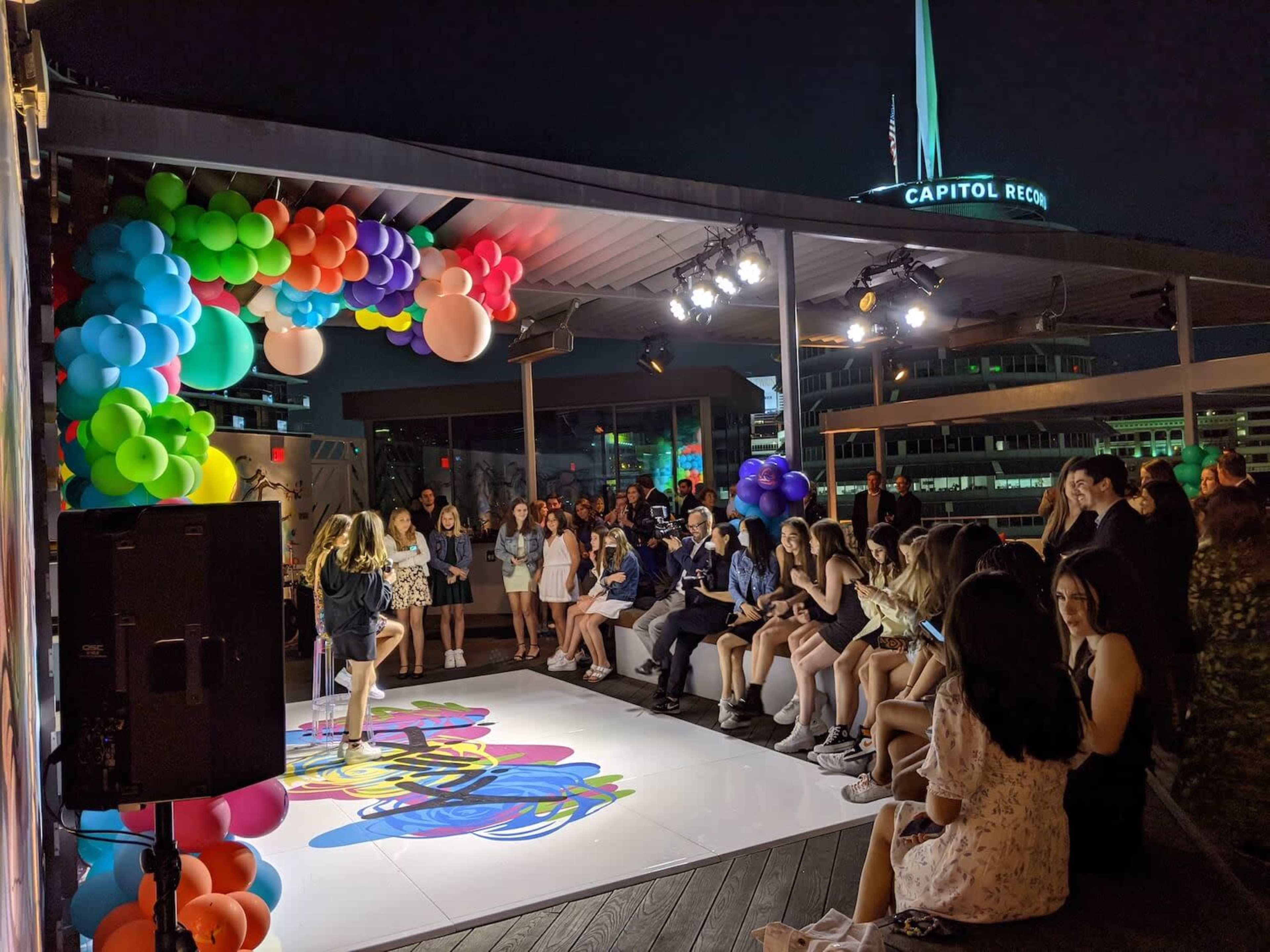 A group of people sits and stands around a brightly lit stage decorated with colorful balloons during an evening event.