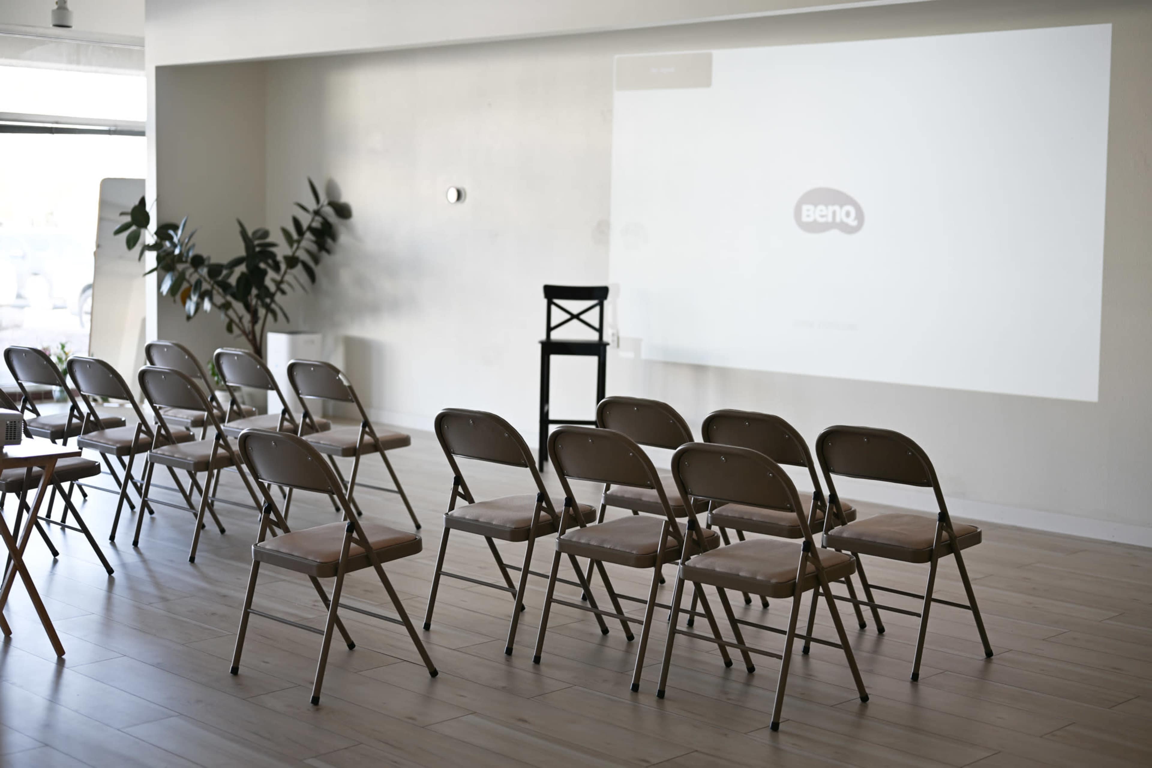A row of folding chairs faces a blank wall with a projector screen displaying the BenQ logo in a simple indoor setting.