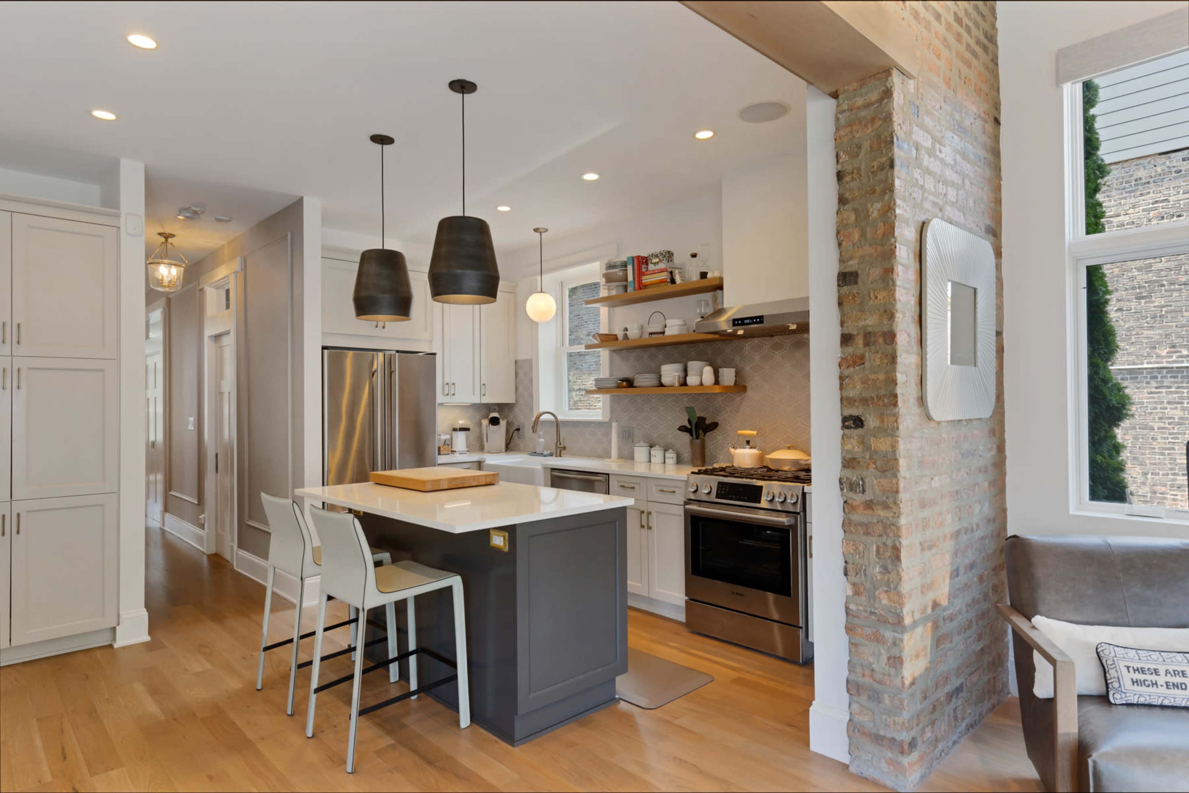 The image shows a modern kitchen with gray cabinets, a large island with bar stools, stainless steel appliances, and exposed brick walls.