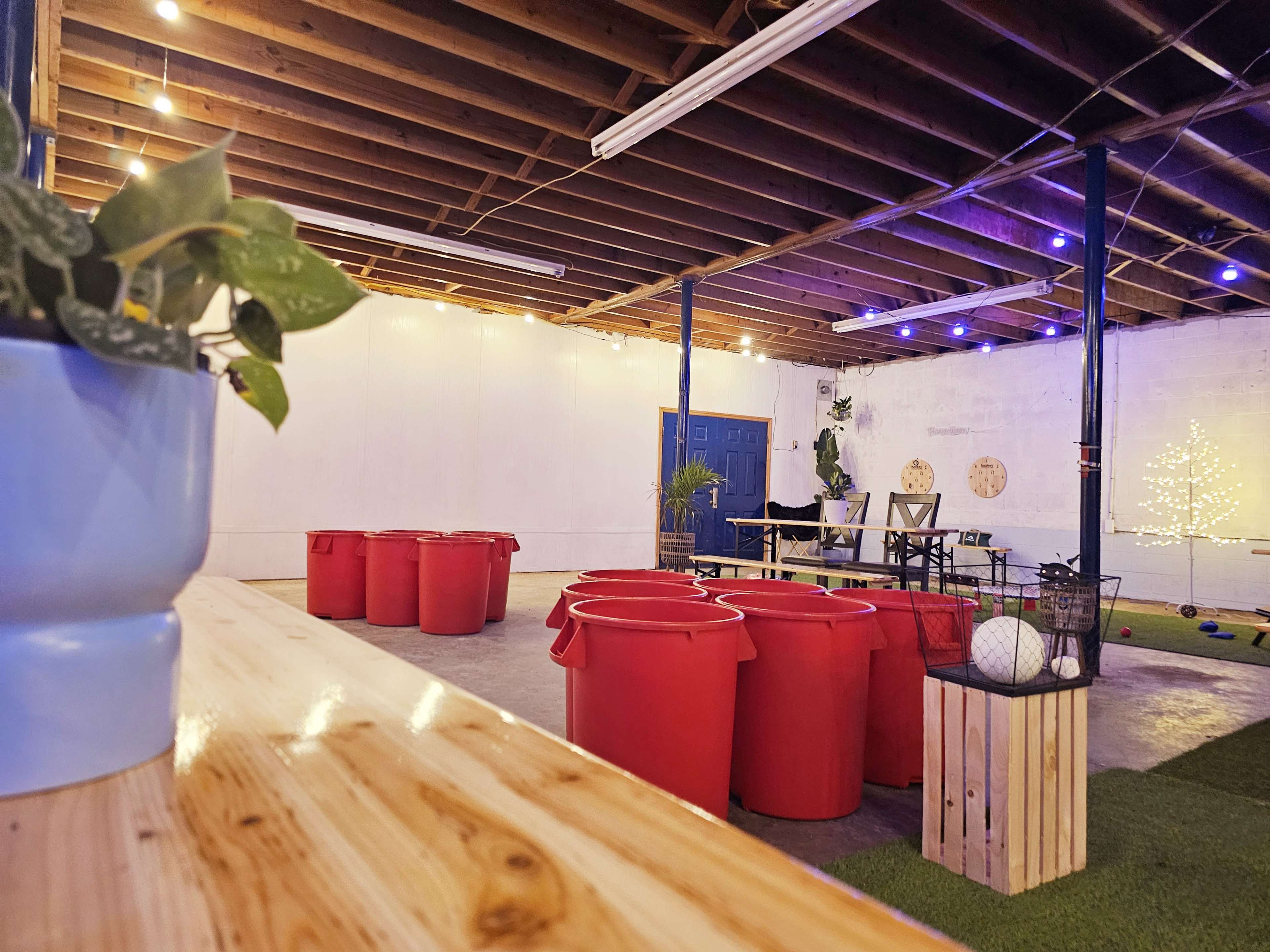 An indoor space with exposed beams, featuring several red barrels, a wooden table with chairs, and decorative lights along the walls.