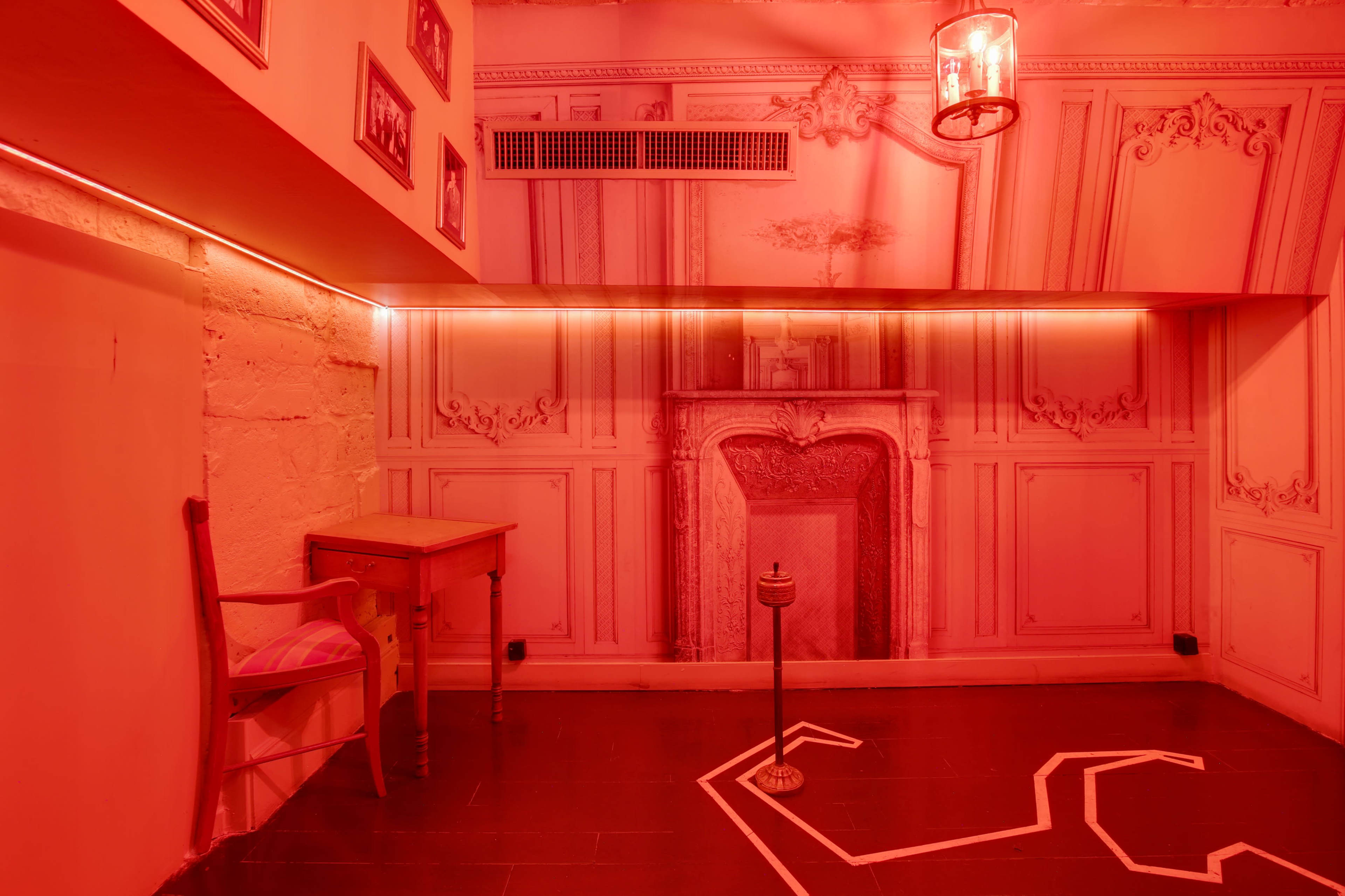 A small room with red lighting, featuring a wall mural of a fireplace, a wooden table, a chair, and a chalk outline on the floor.