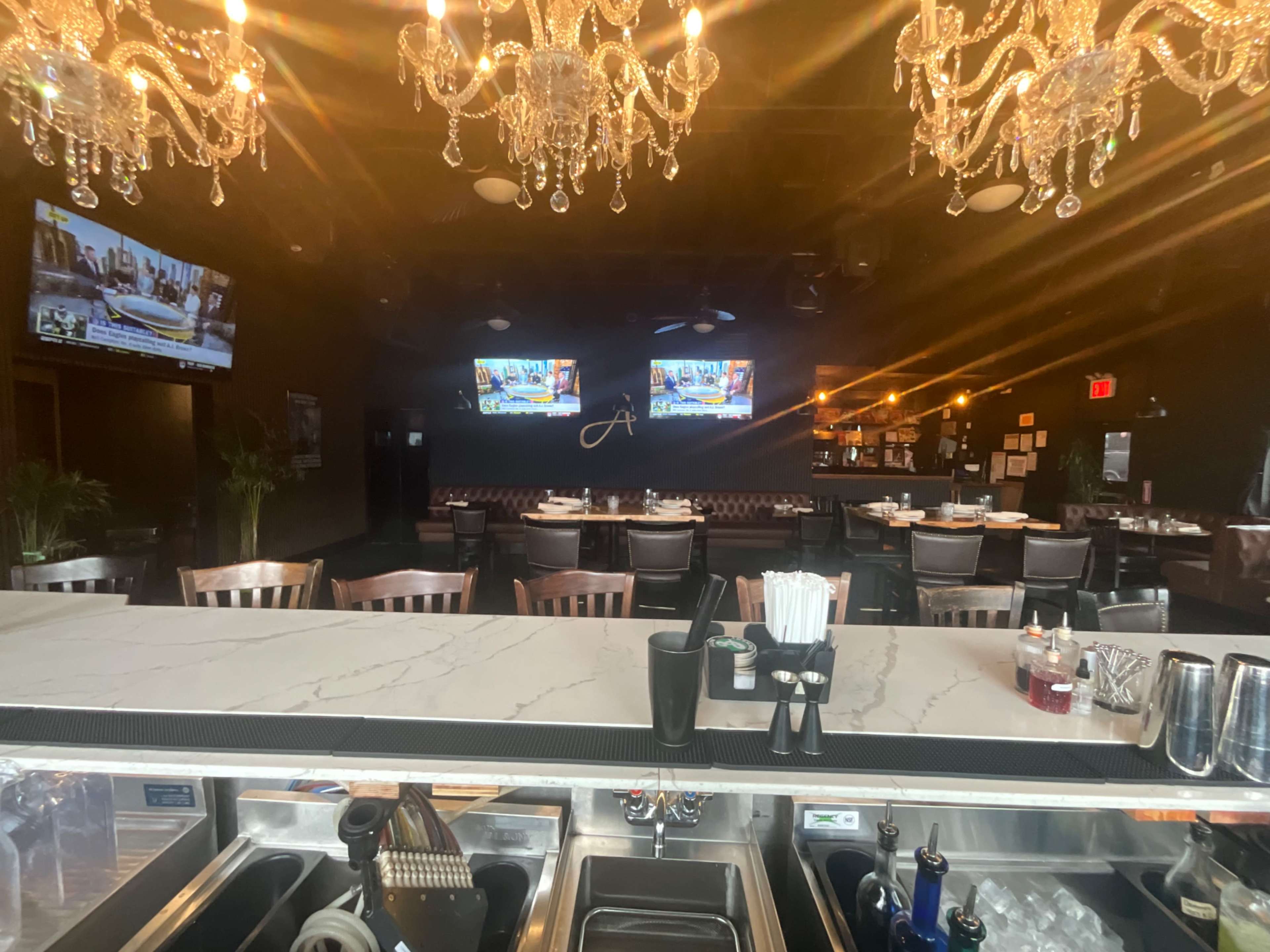 The image shows the interior of a restaurant featuring a bar area with a marble countertop, crystal chandeliers, and tables set for dining, along with two televisions mounted on the wall.