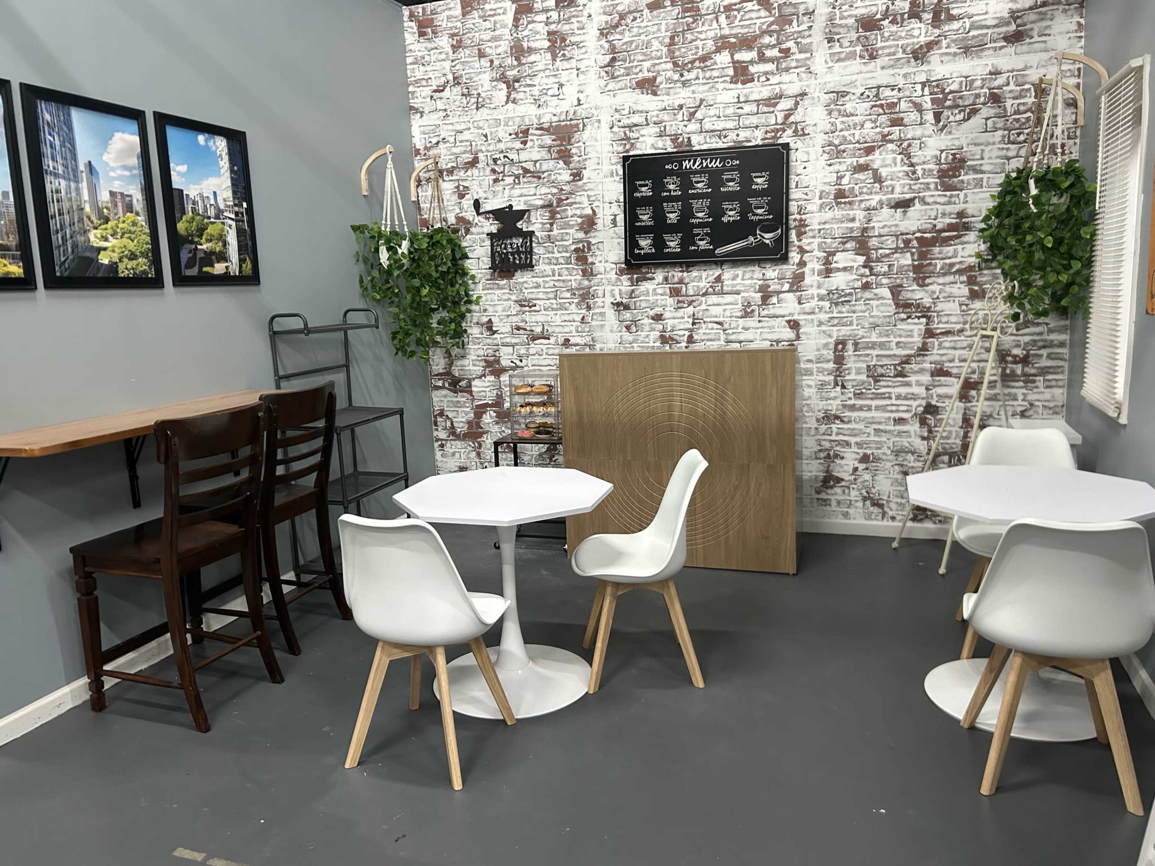 The image shows a small cafe interior with white tables and chairs, a wooden counter, and brick-patterned walls adorned with pictures and greenery.