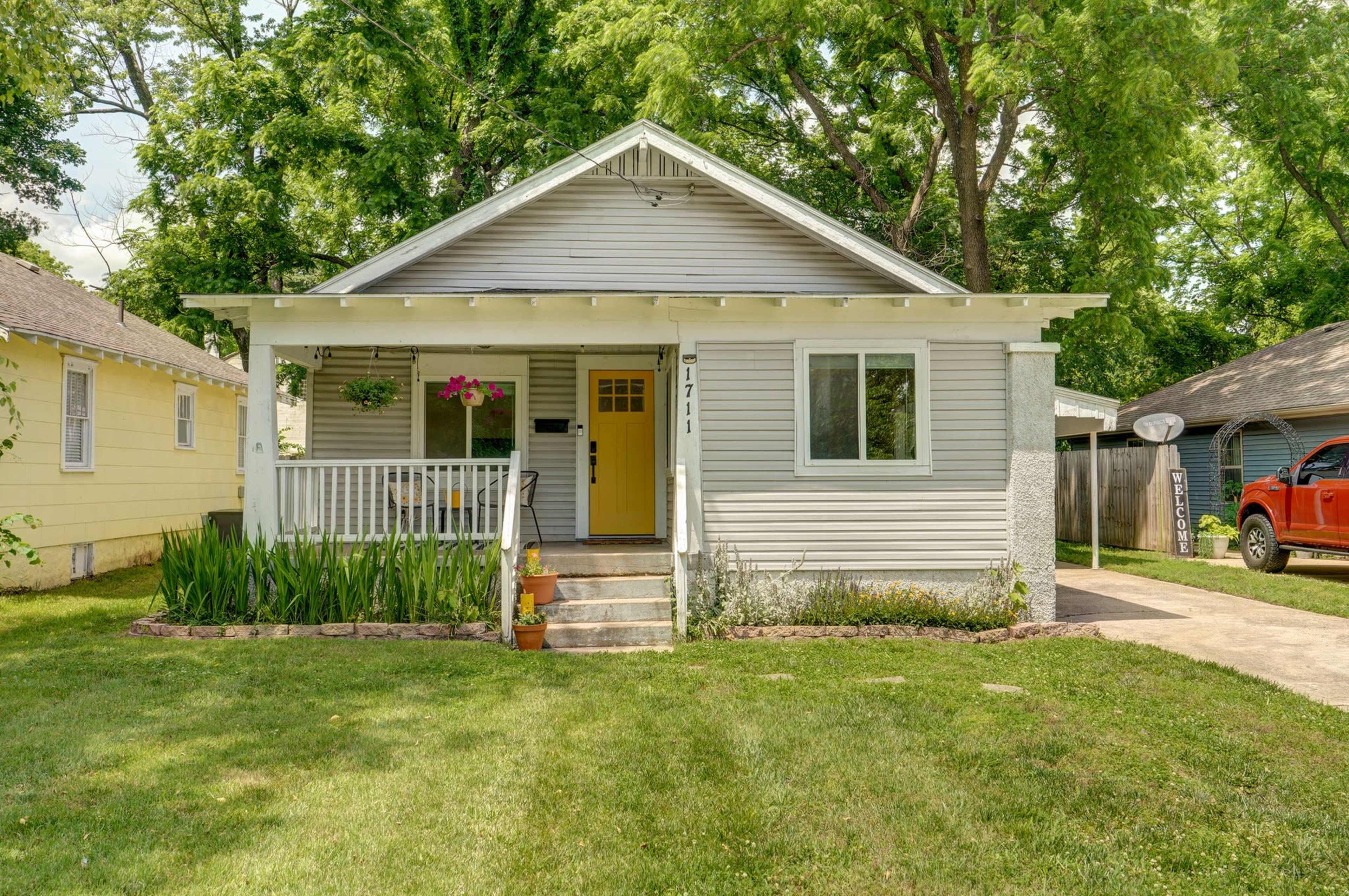 A single-story, light gray house with a front porch, surrounded by green grass and trees, has a driveway leading to a parked red truck.