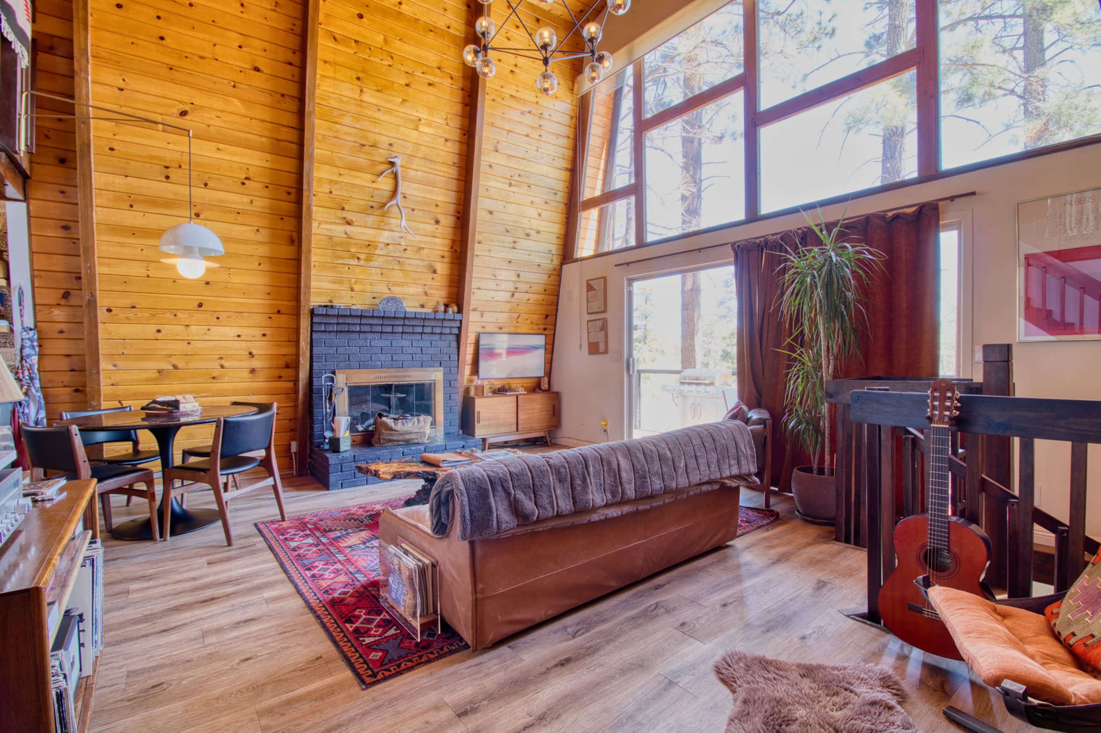 The image features a cozy living room with a wooden A-frame structure, a fireplace, a dining area, and large windows allowing natural light to enter.