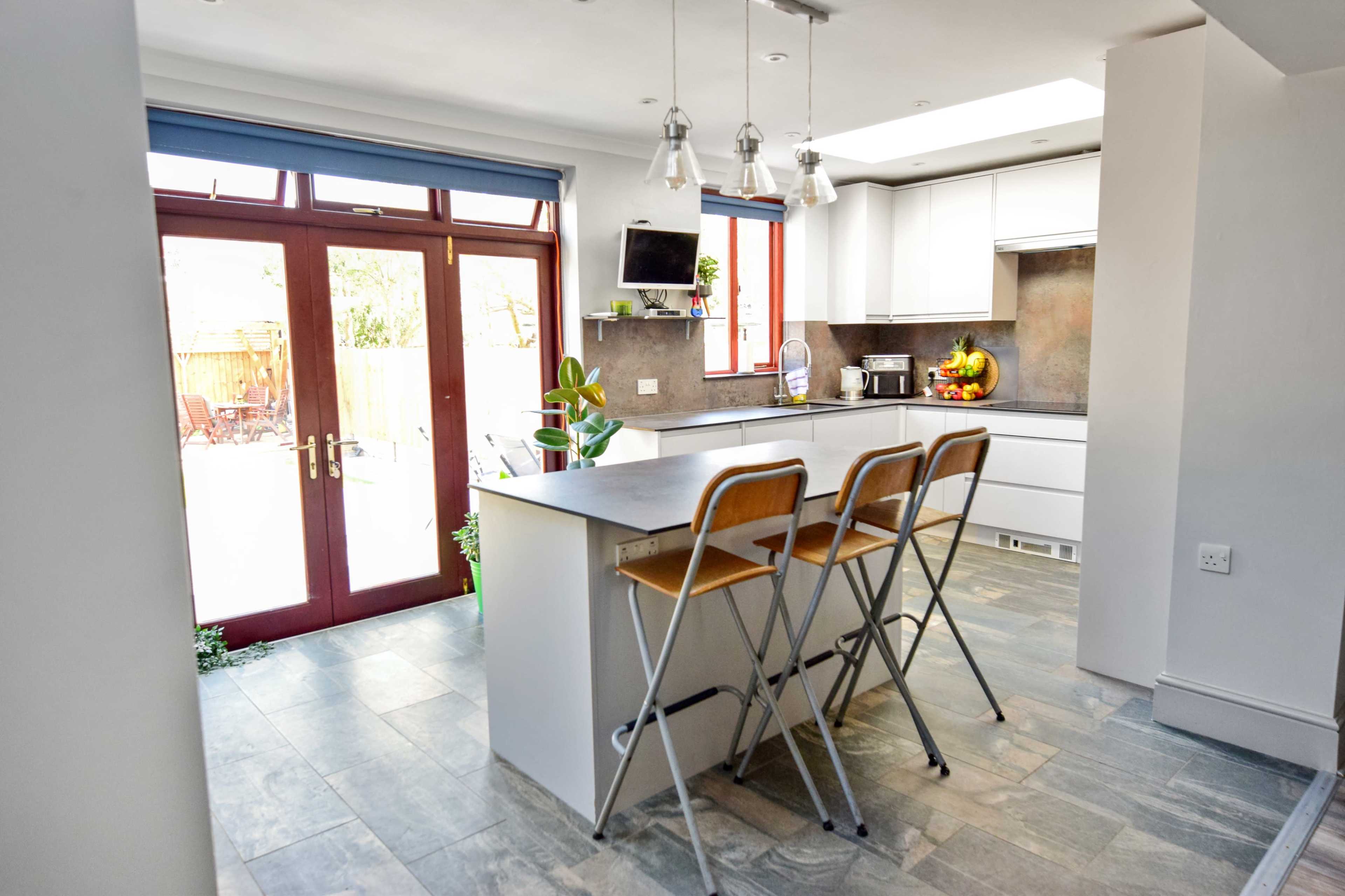 A modern kitchen with a central island and three wooden stools, featuring large windows and a door that leads to an outdoor space.