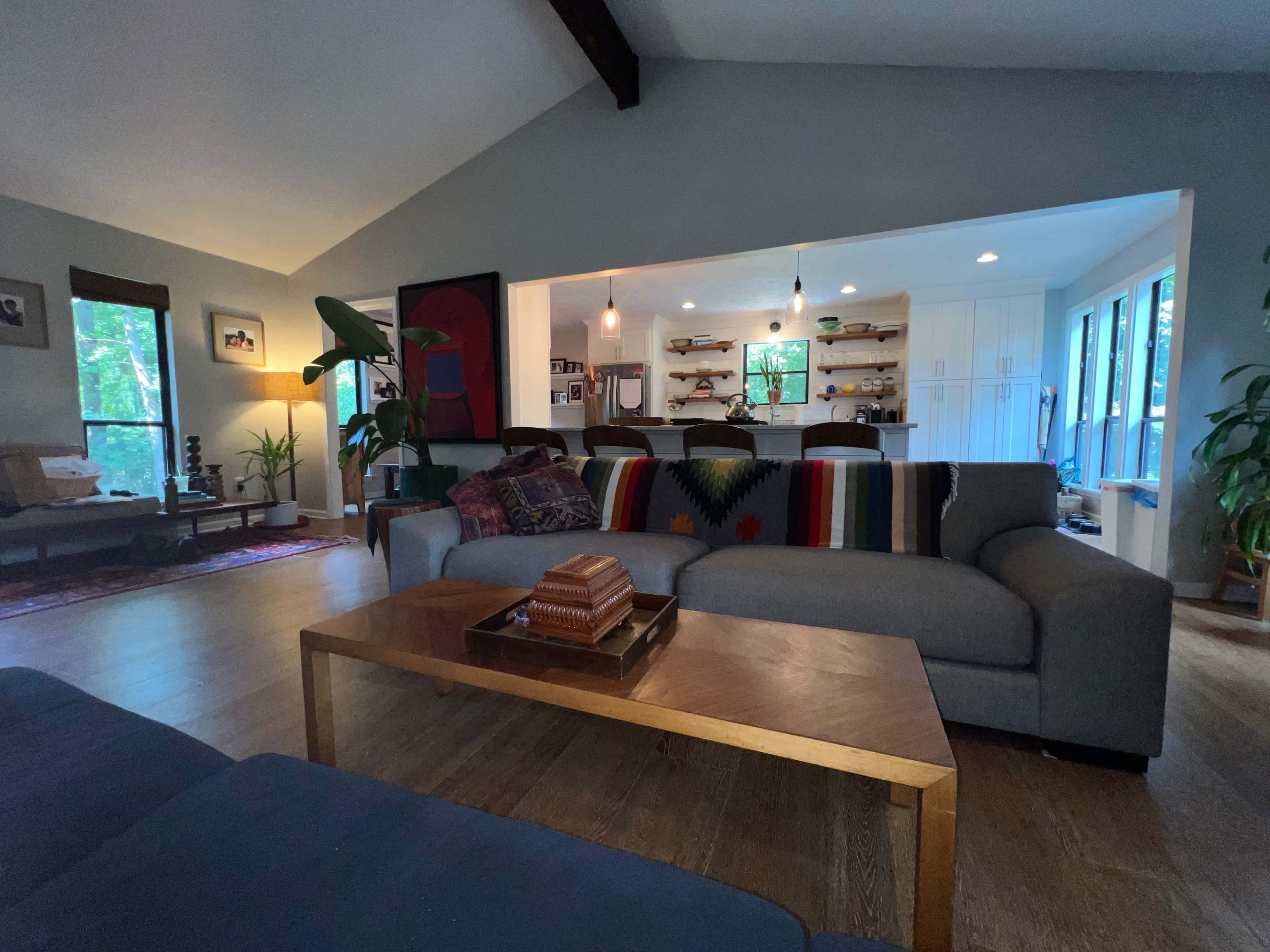 A living room with a sofa, a wooden coffee table, and an open kitchen in the background, featuring shelves and seating.