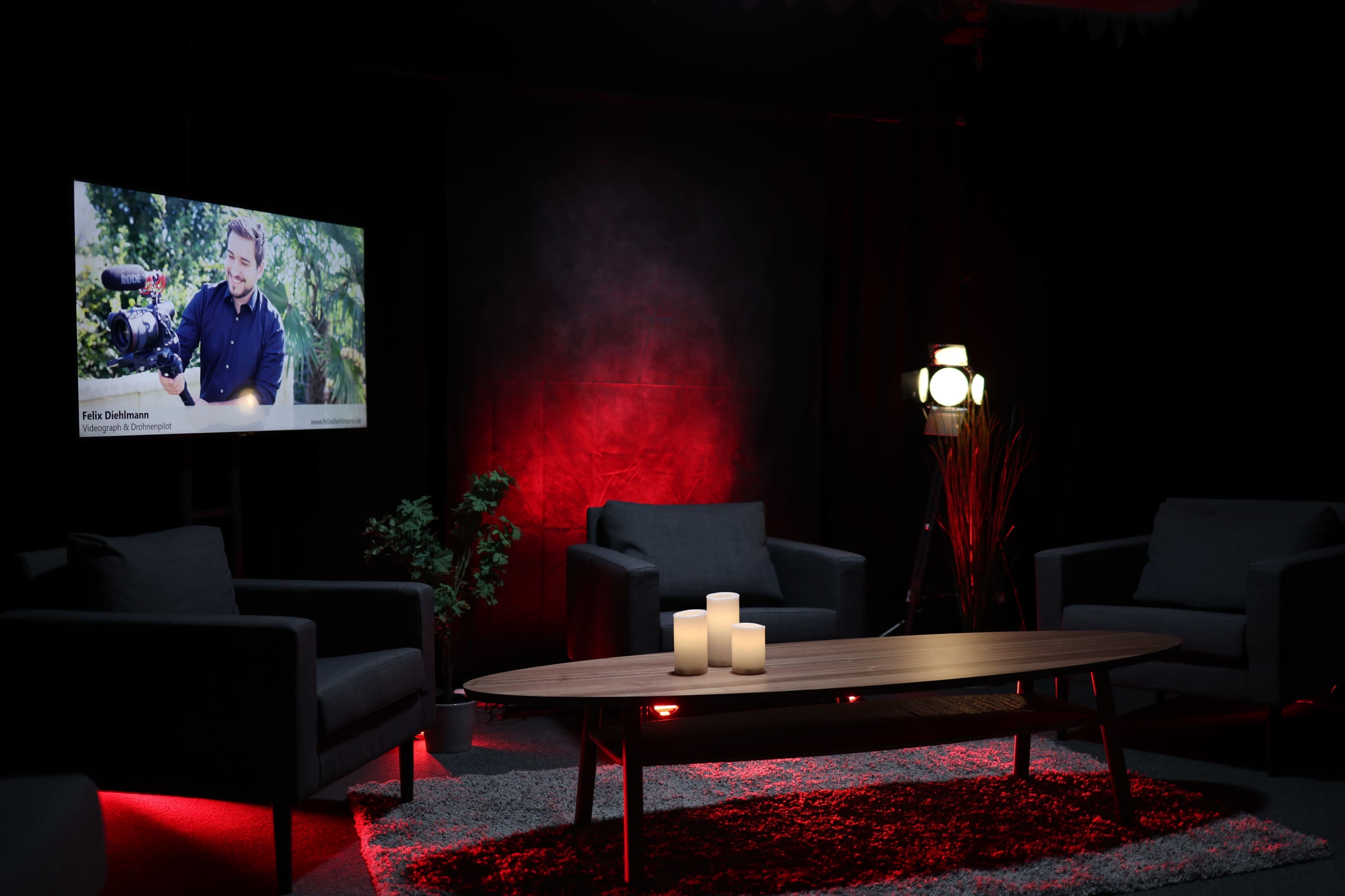 The scene features a modern, dimly lit studio setup with two black sofas facing a wooden coffee table, three candles on the table, and a screen displaying a man in a garden.