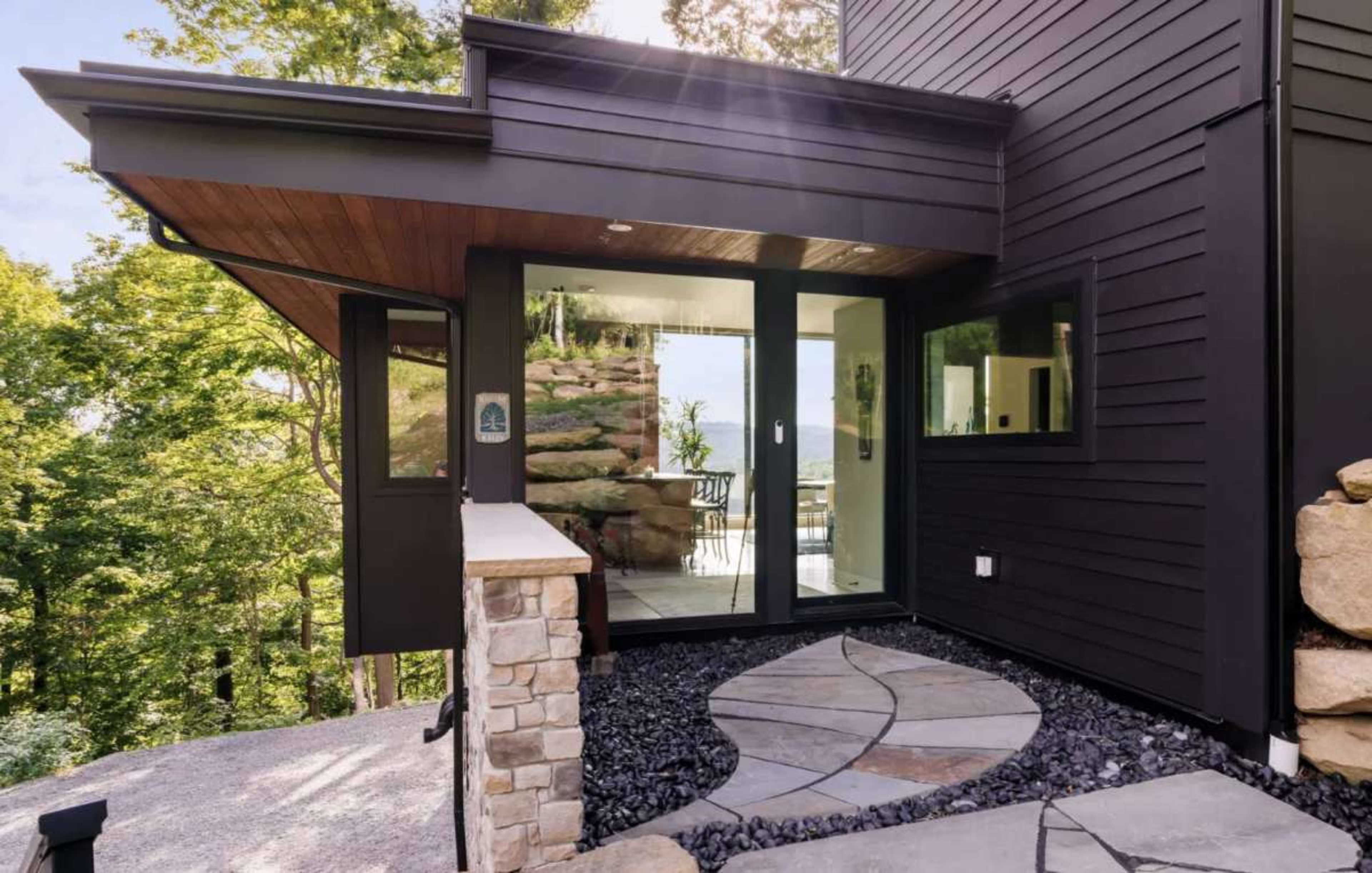 The image shows a modern home entrance with a stone pathway leading to large glass doors surrounded by natural greenery.