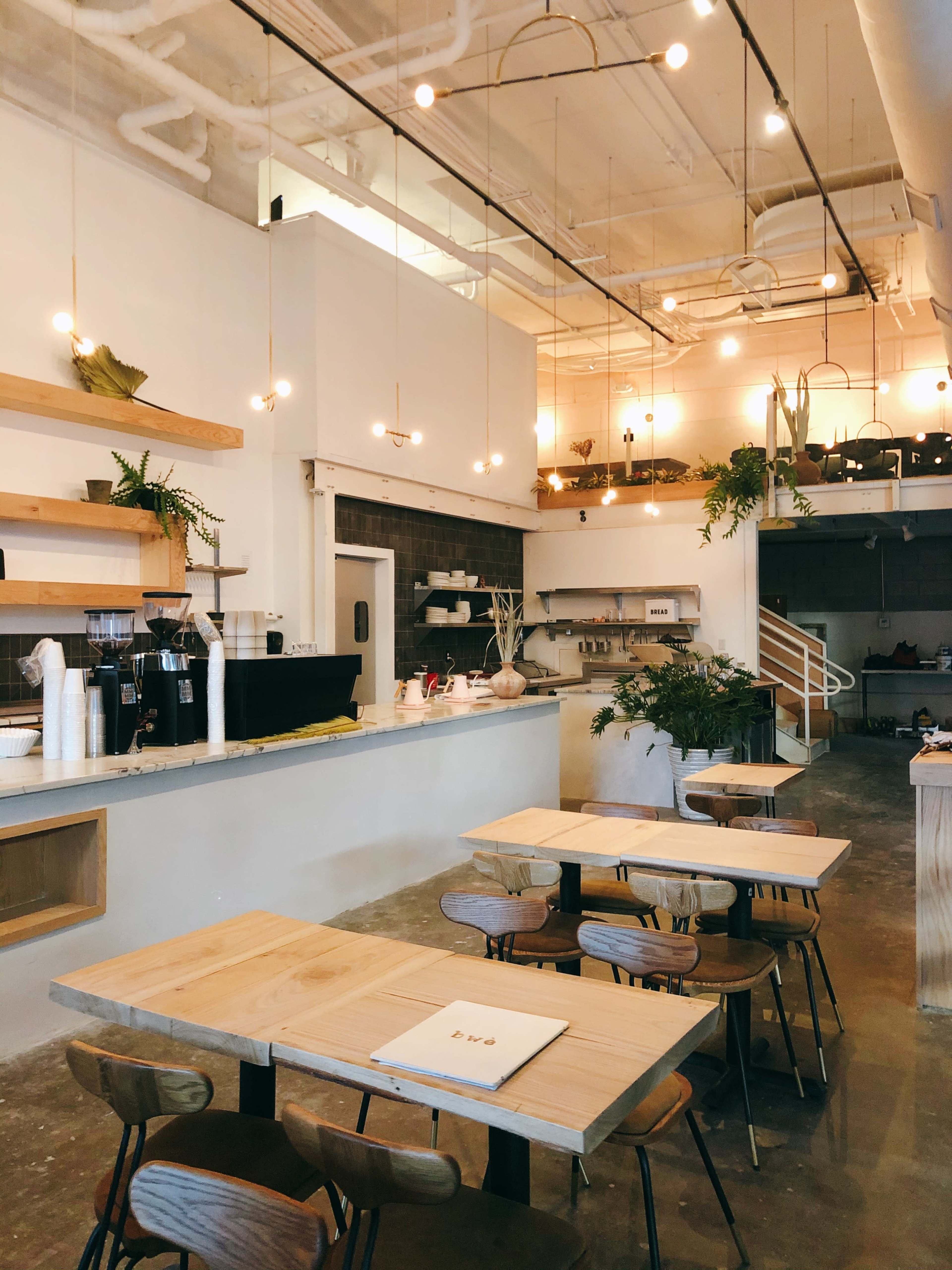 The image shows a modern café interior with wooden tables, pendant lighting, and a minimalist counter area featuring plants and kitchen equipment.