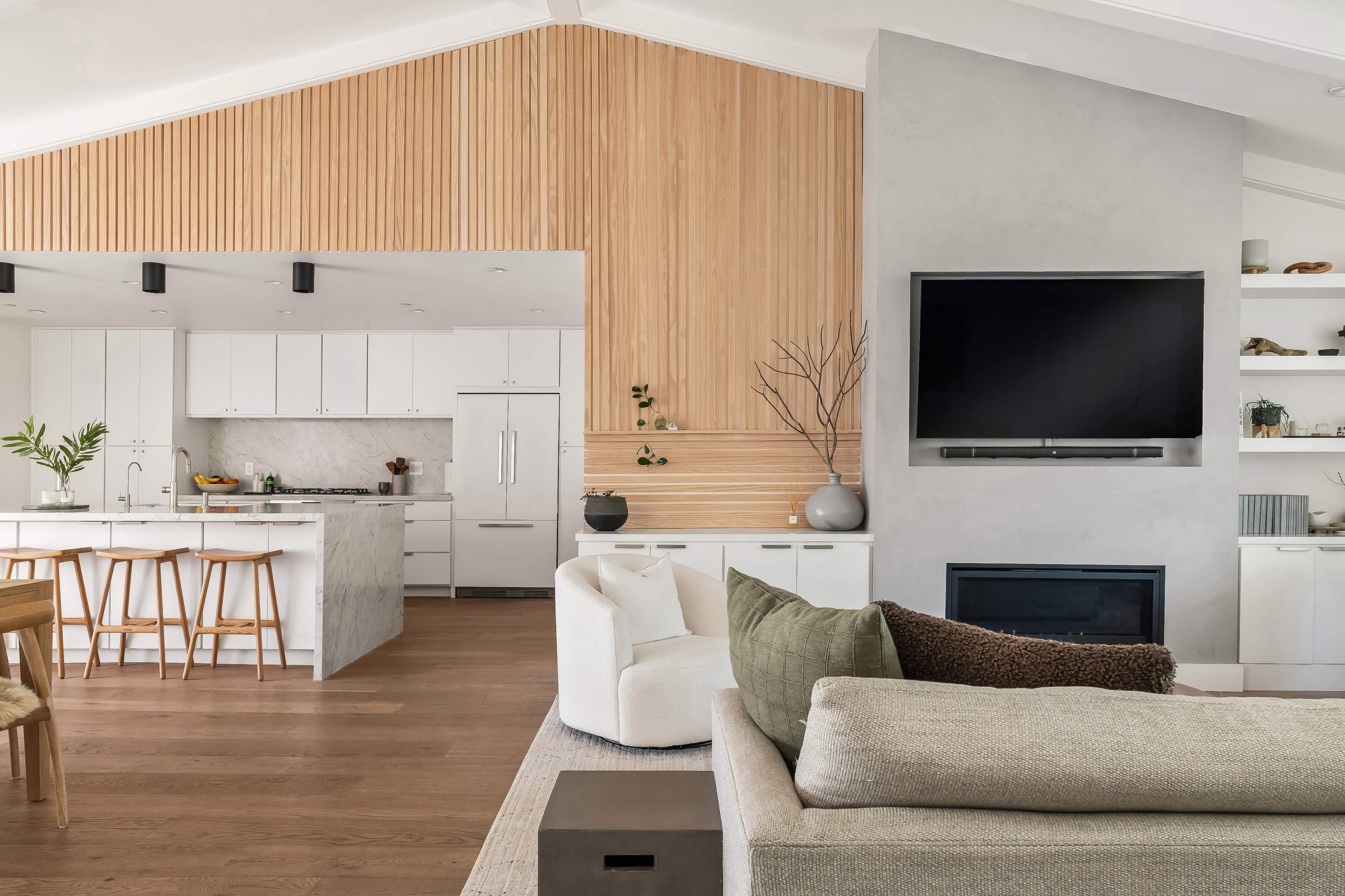 The image shows a modern living space featuring an open floor plan with a kitchen that has white cabinets and a marble island, alongside a cozy seating area with a large television mounted on a gray wall.