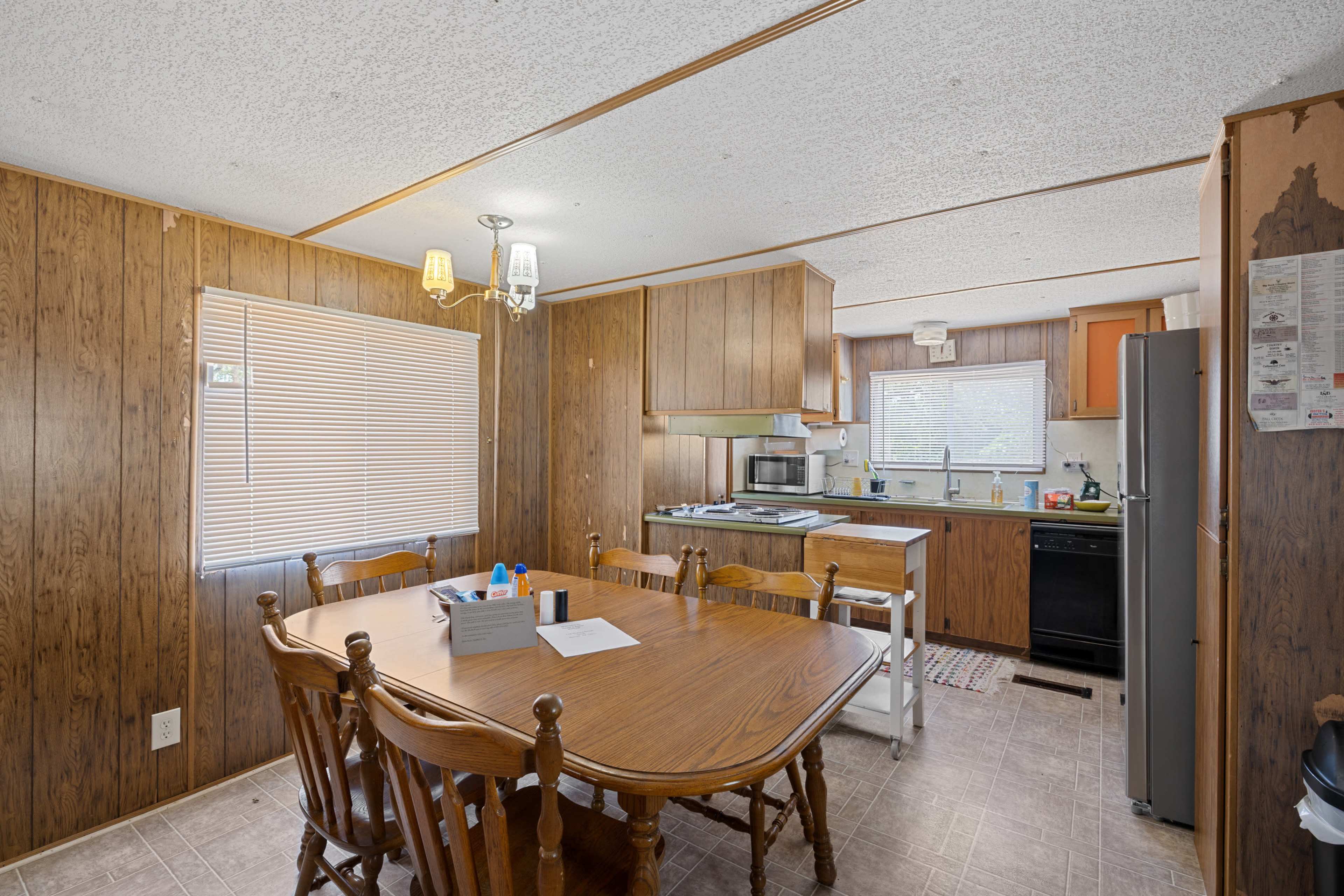 The image shows a modest kitchen and dining area, featuring a wooden table with chairs, cabinets, and appliances against a brown panelled wall.