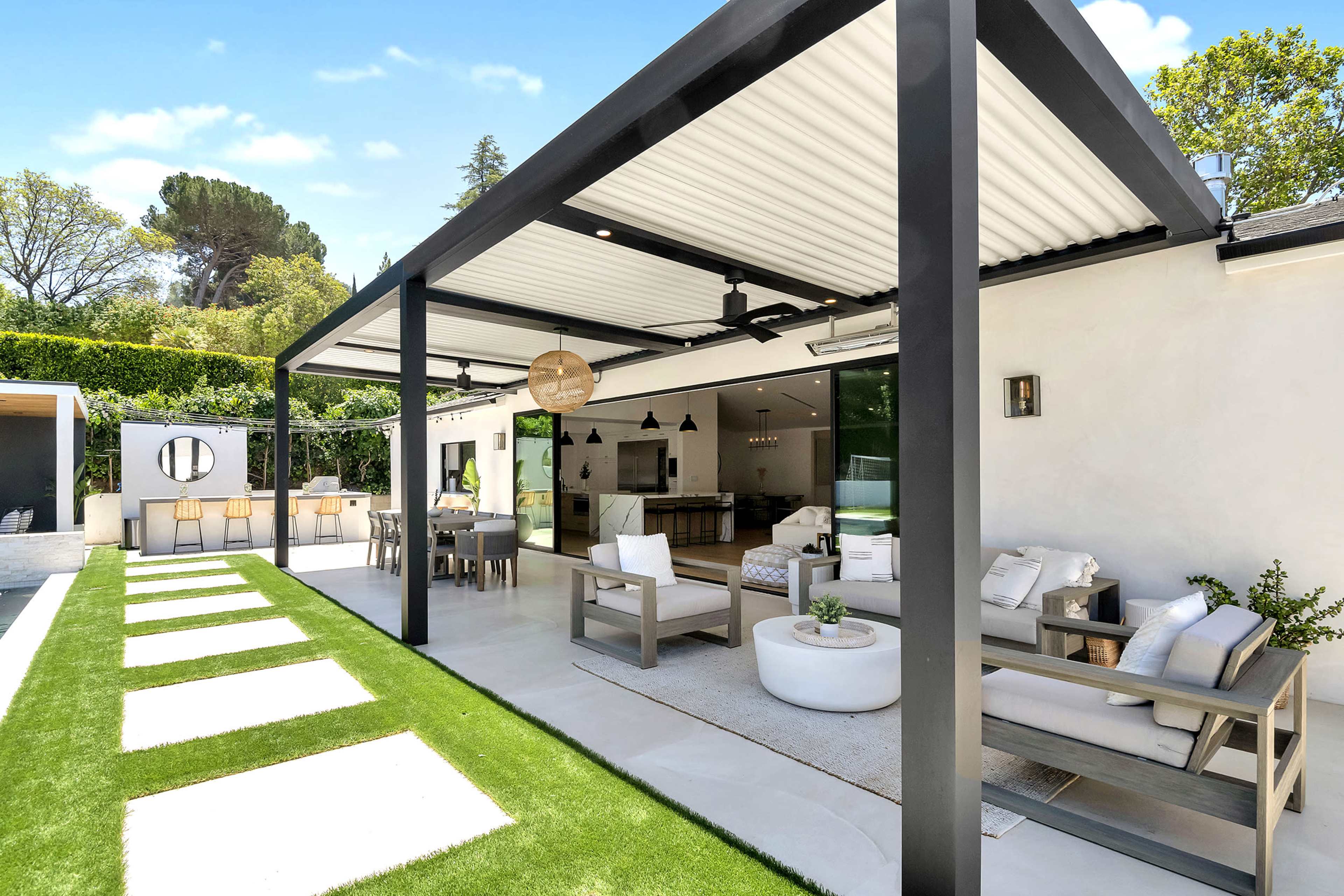 A modern outdoor patio features a covered seating area with a dining space, surrounded by neatly arranged stone path and lush green grass.