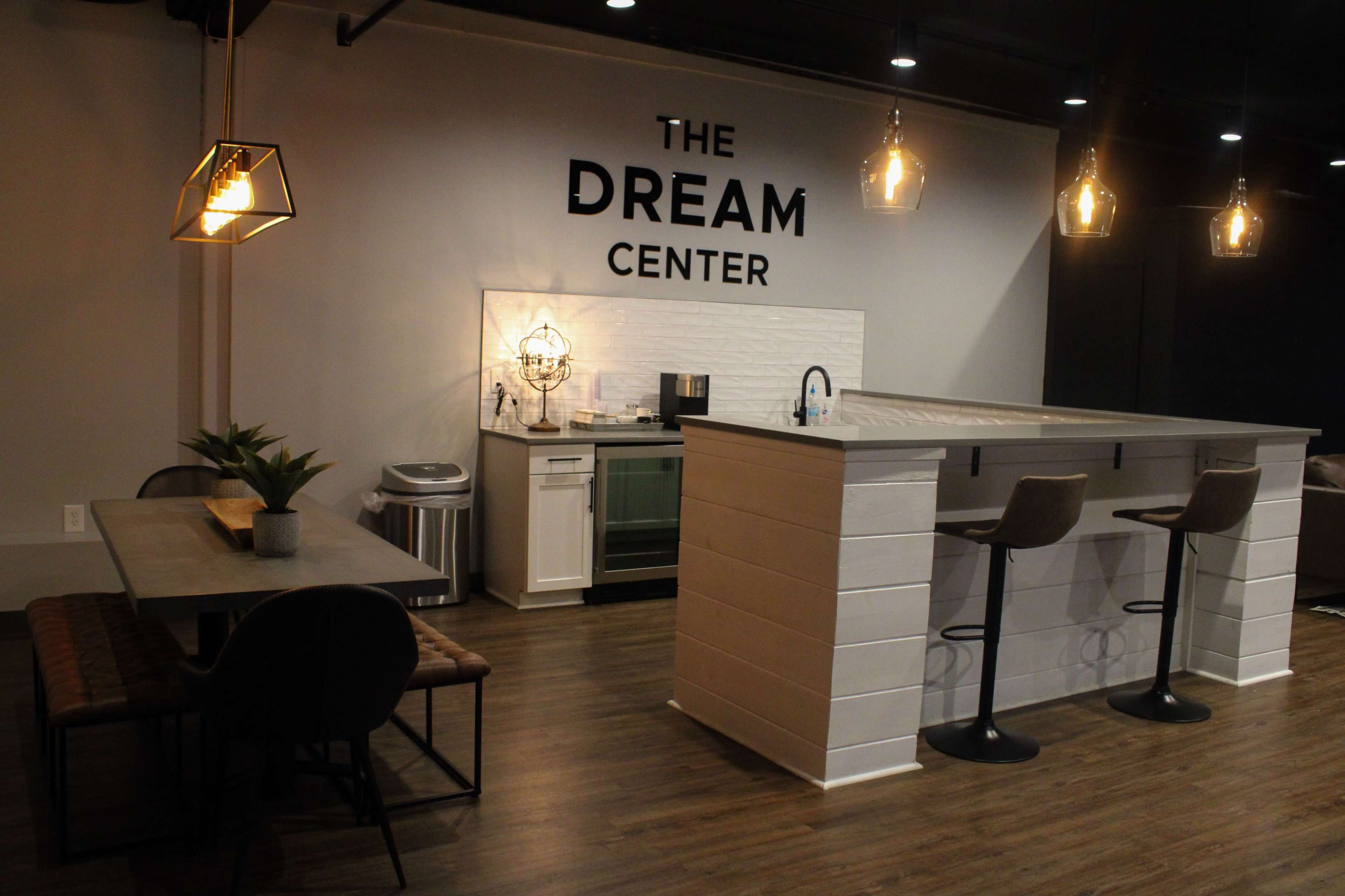 A modern kitchen area with a bar and seating, alongside a dining table, featuring a sign that reads "THE DREAM CENTER" on the wall.