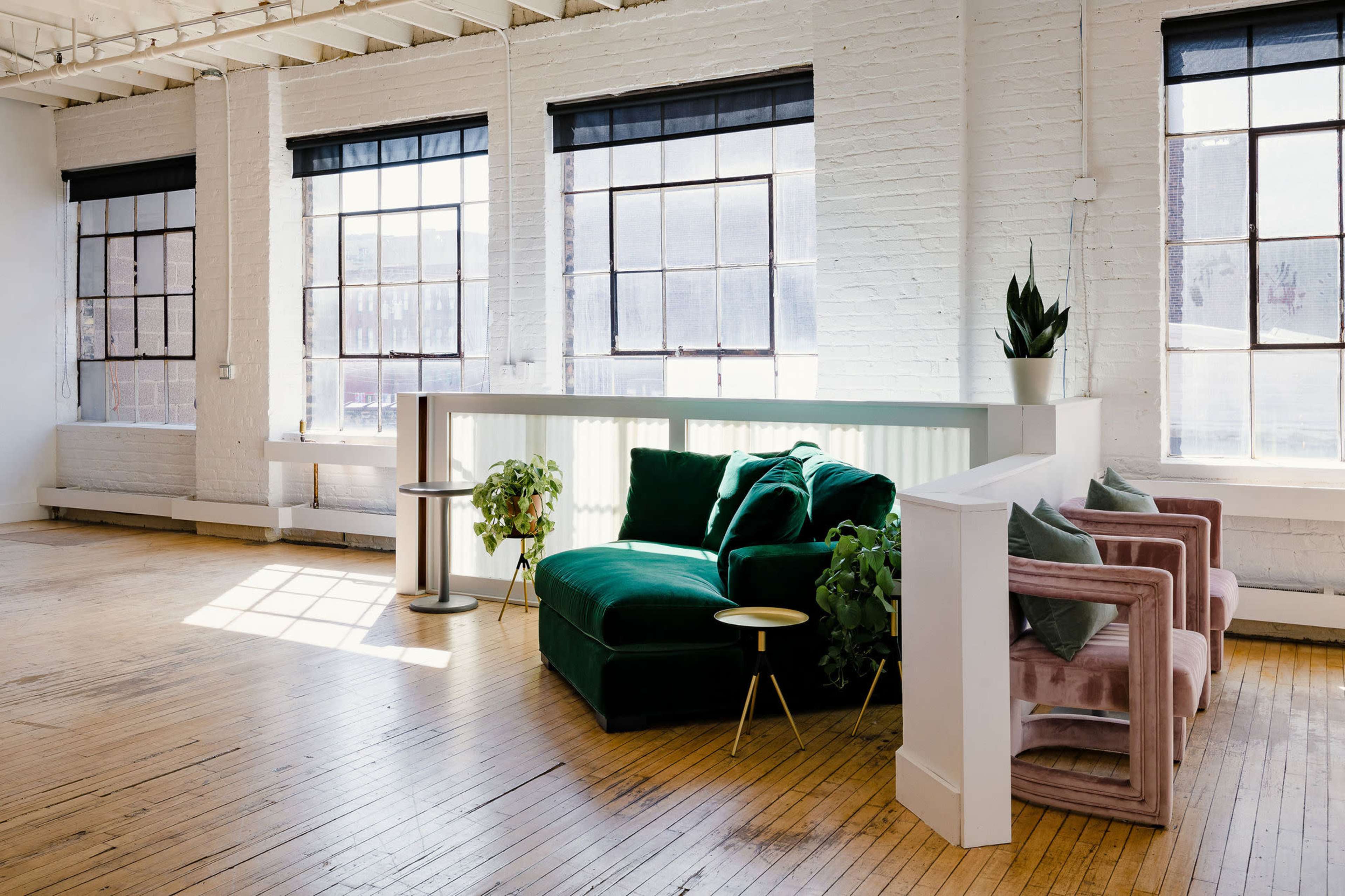 A spacious interior with hardwood floors, large windows, and a green velvet couch along with pink armchairs and potted plants.