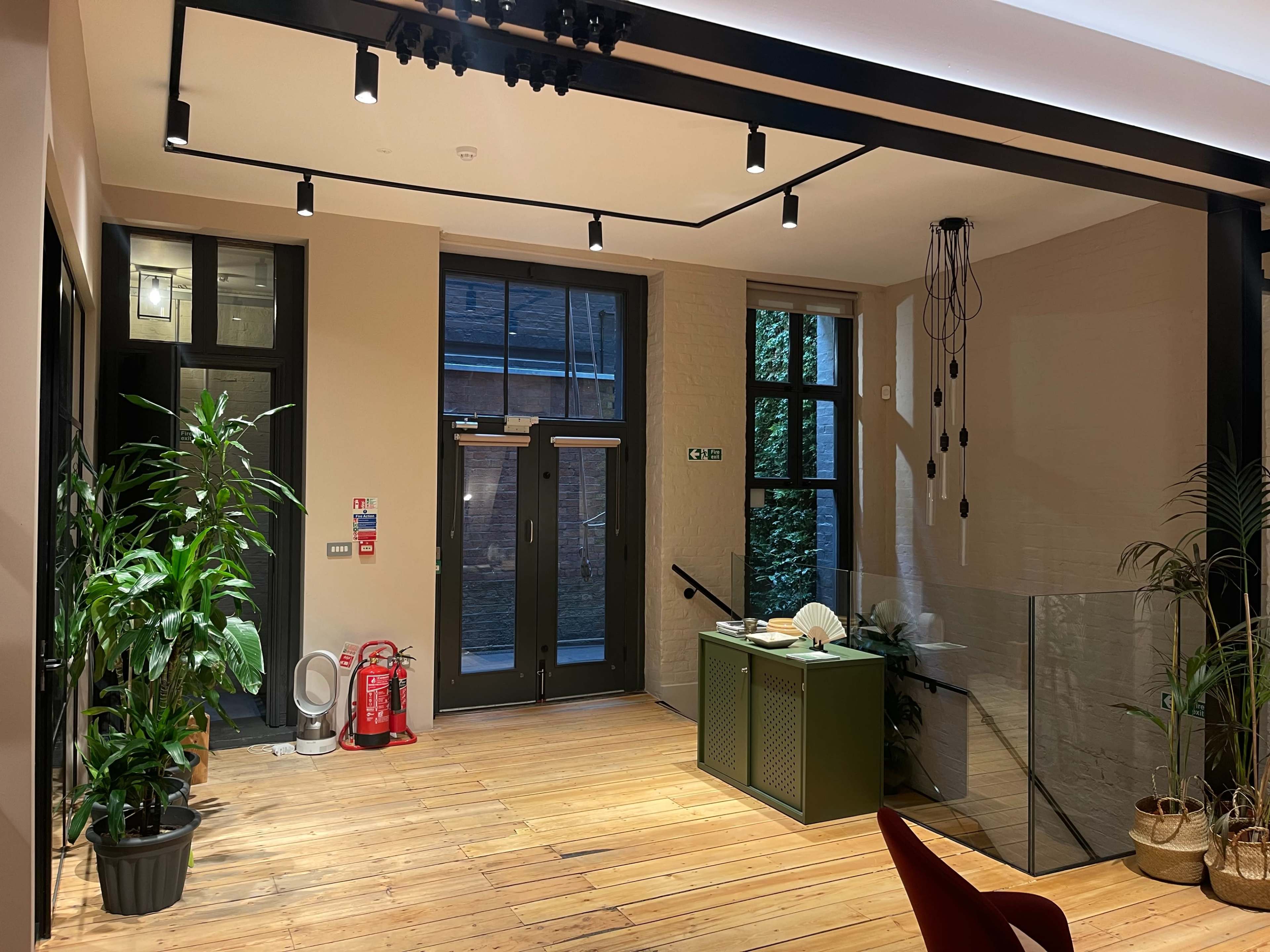 The interior of a modern space features wooden flooring, potted plants, and a reception area with a green desk, flanked by large windows and visible exit doors.