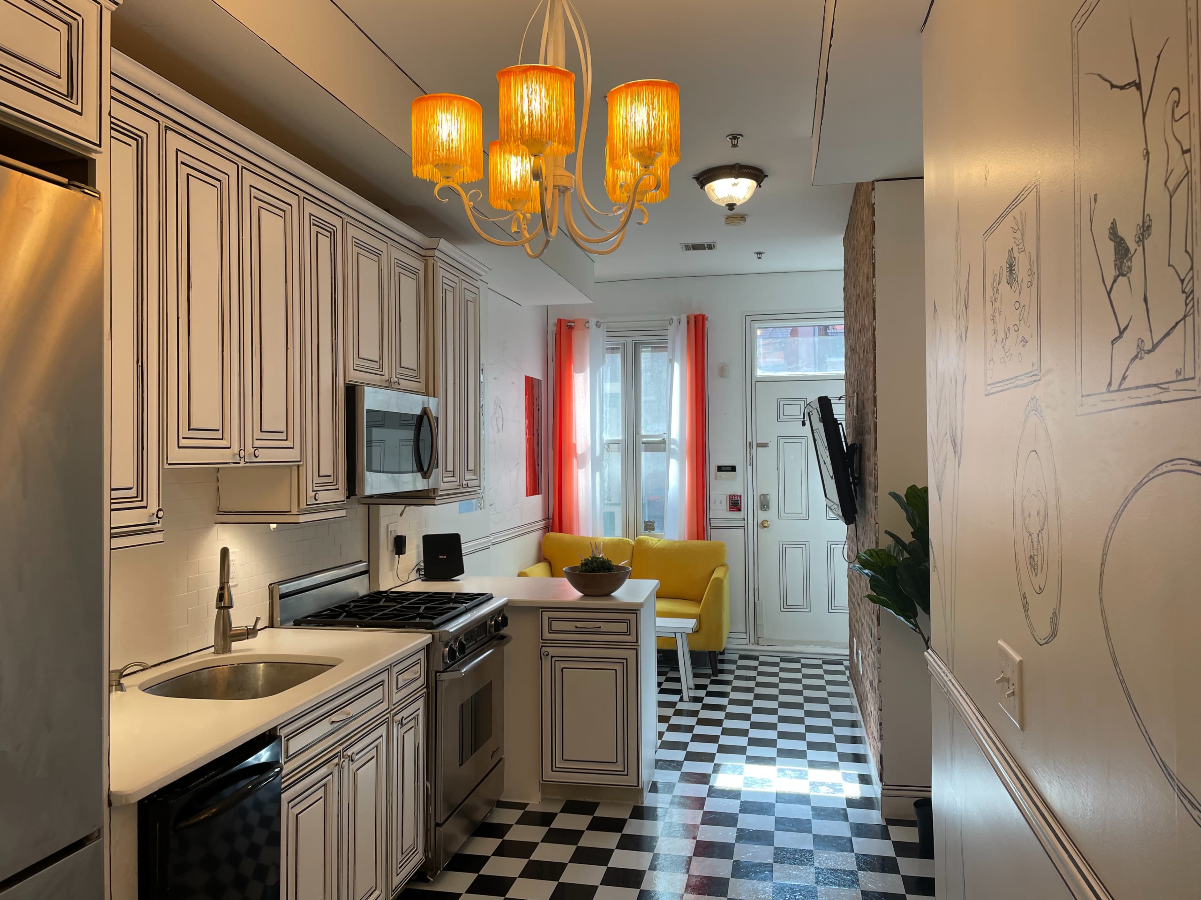 A kitchen with white cabinetry, a stainless steel refrigerator, and a checkered black-and-white floor, leading into a bright living area with a yellow sofa.