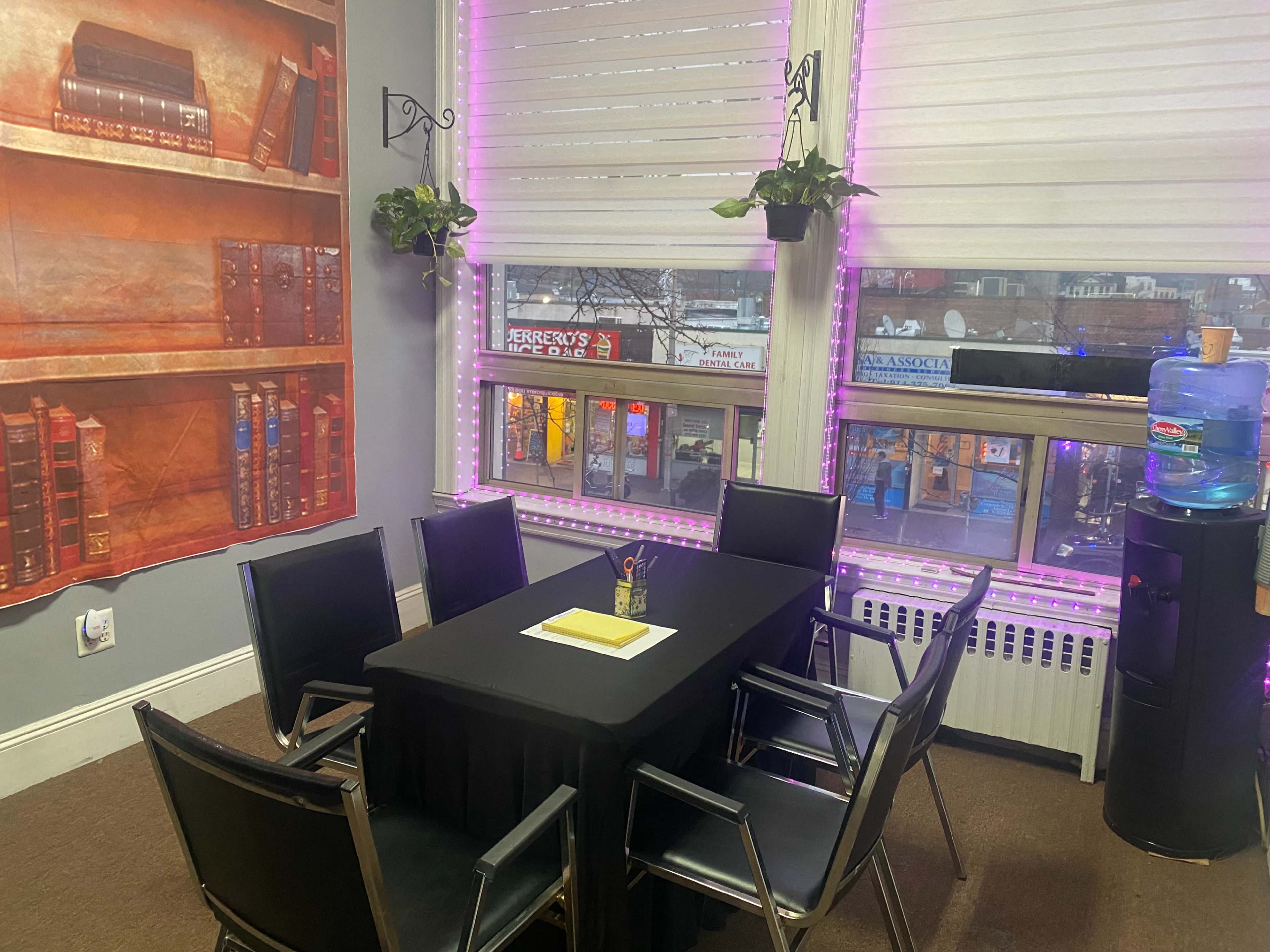 A small meeting room with a black table surrounded by six chairs, a water cooler, and bookshelves with a backdrop of a street visible through large windows.