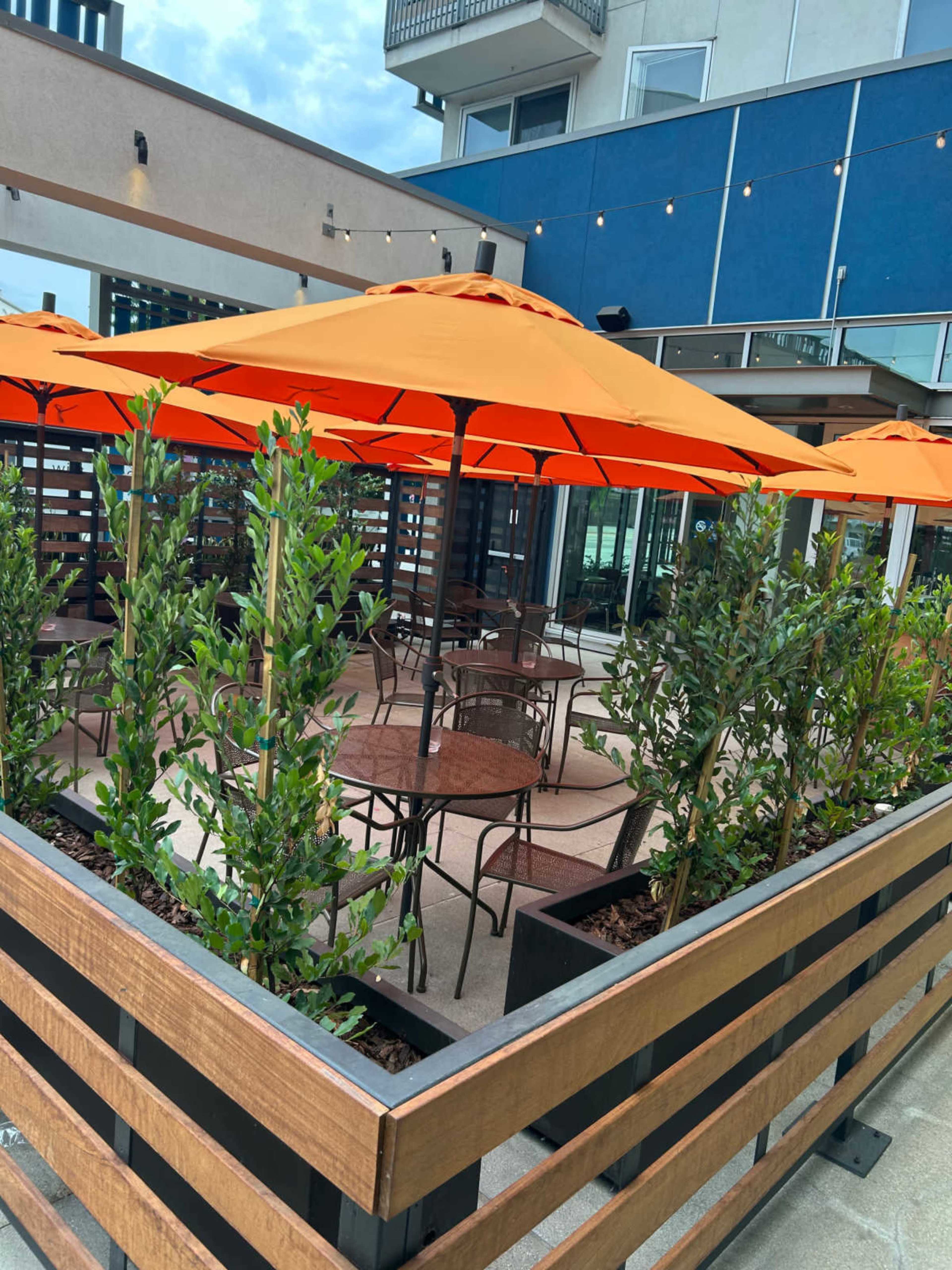 The image shows a patio area of a restaurant, featuring tables and chairs surrounded by planters and orange umbrellas.