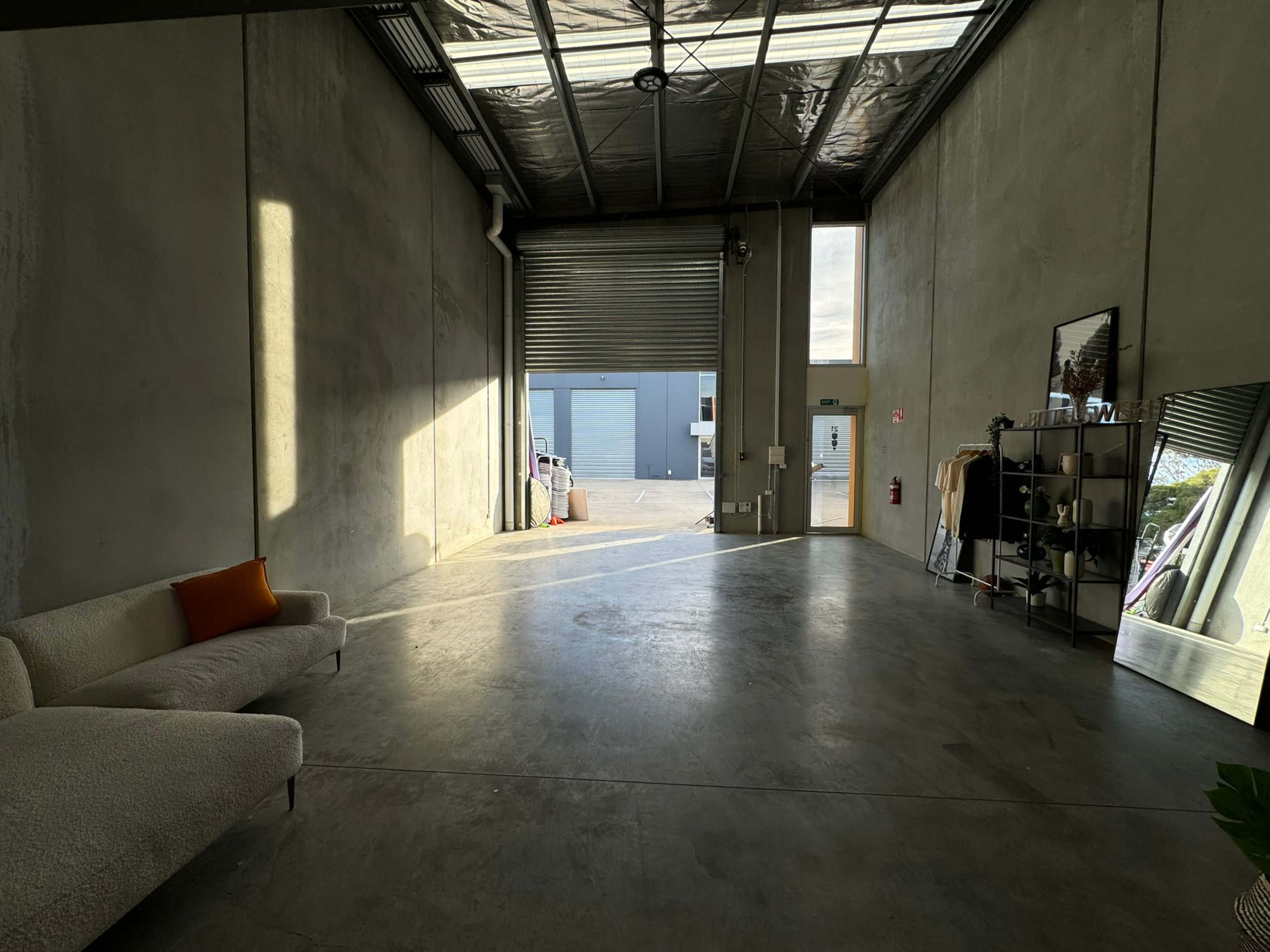A spacious industrial interior with concrete walls and floors, featuring a sofa, a mirror, and an open garage door leading to a loading area.