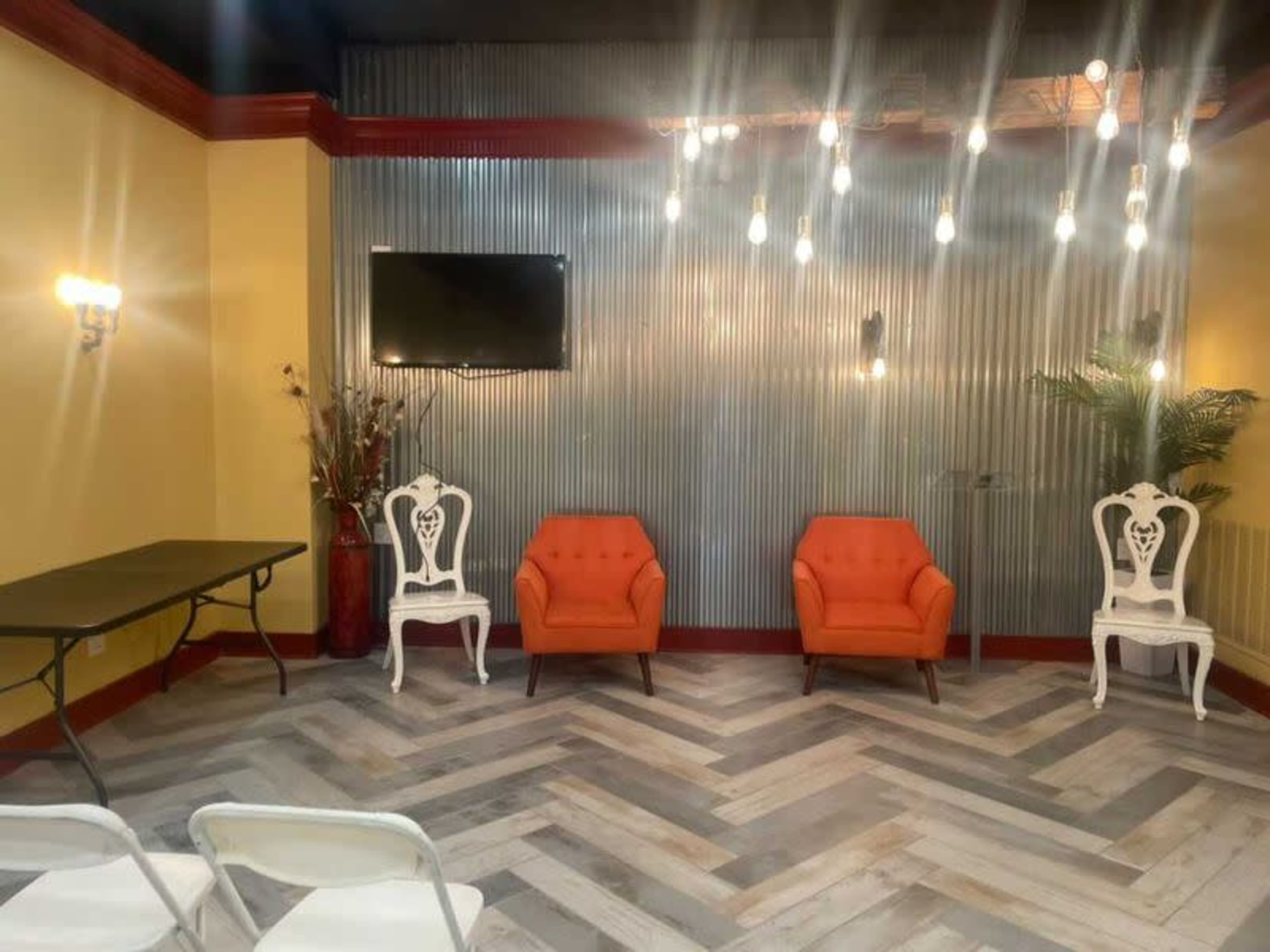 A room with two orange chairs, white chairs, a black table, a television on the wall, and exposed lights hanging from the ceiling.