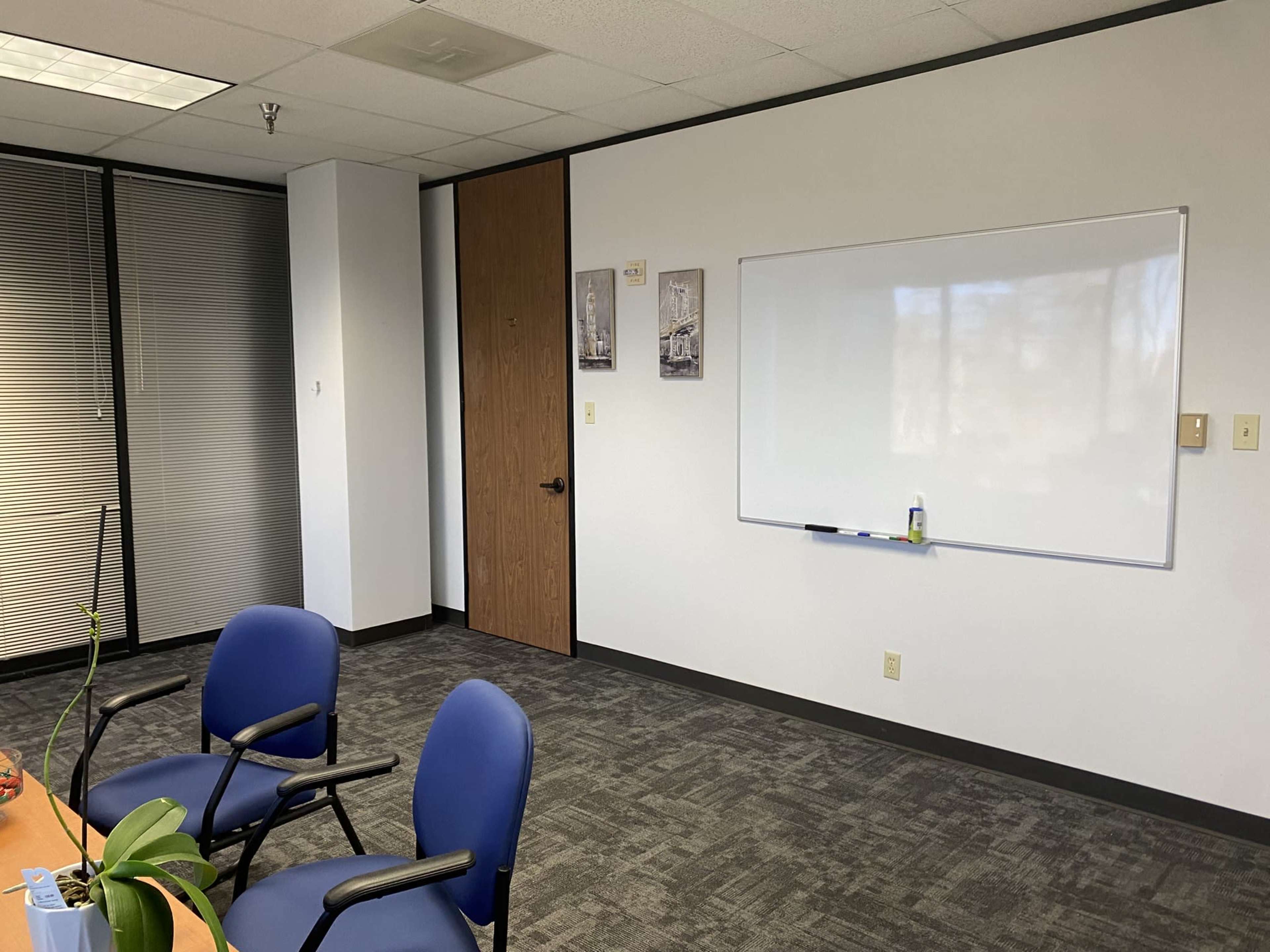 The image shows an office space featuring a round table with blue chairs, a whiteboard mounted on the wall, and a door leading to another area.