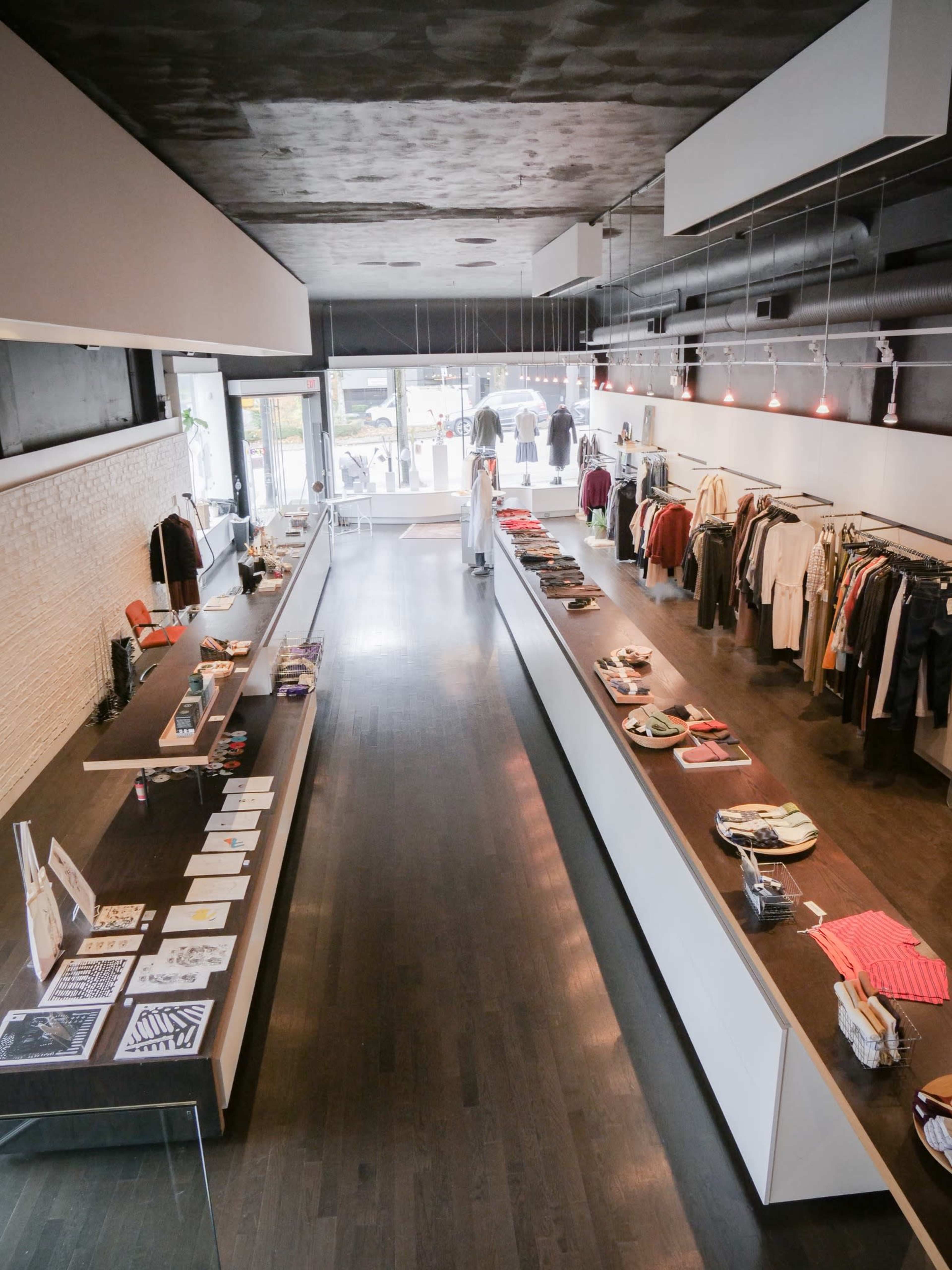 A modern retail clothing store with long display tables featuring apparel and accessories, and a large window displaying the street outside.