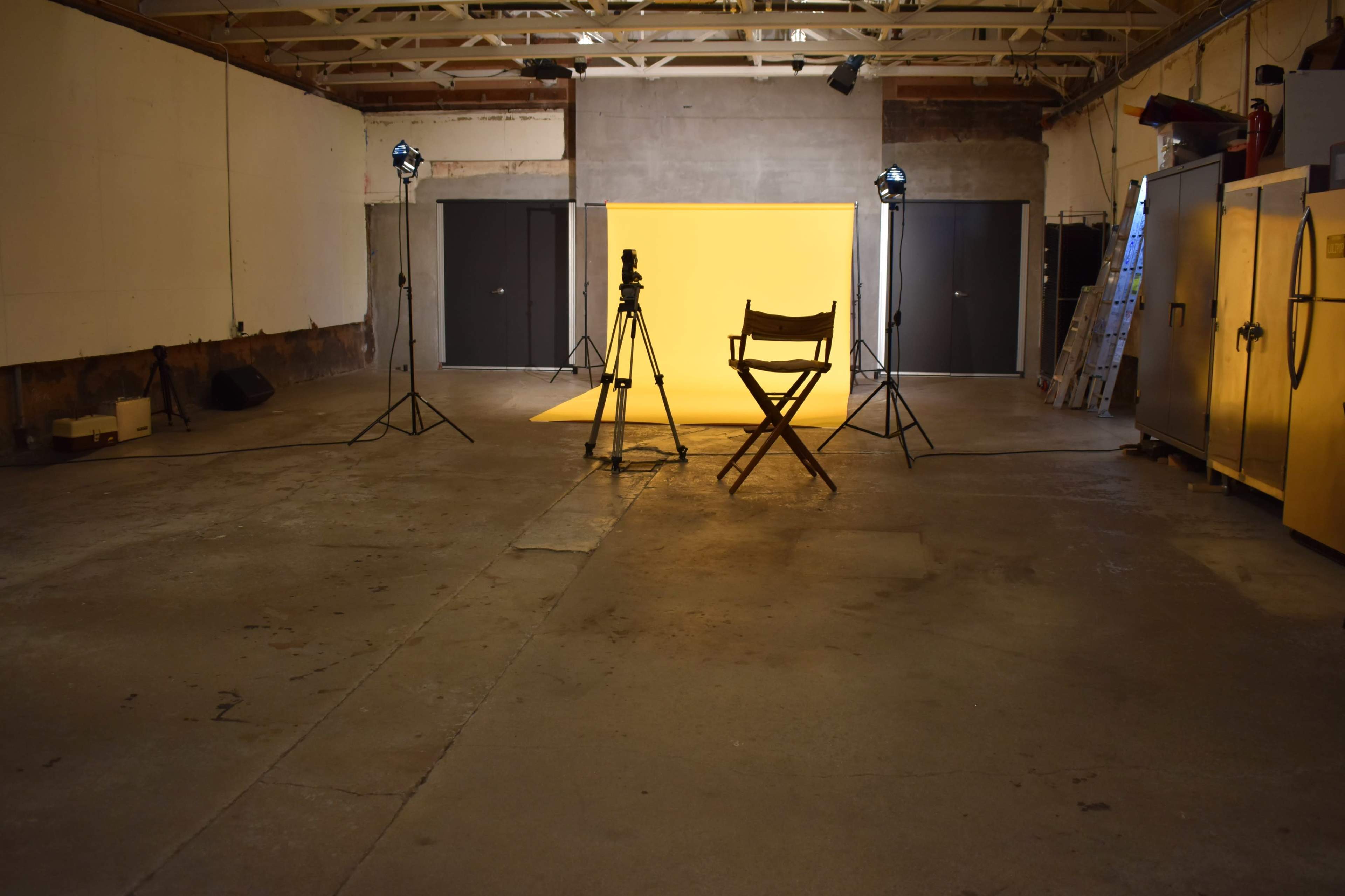 The image depicts a spacious studio with a yellow backdrop, a director's chair, and two cameras on tripods.