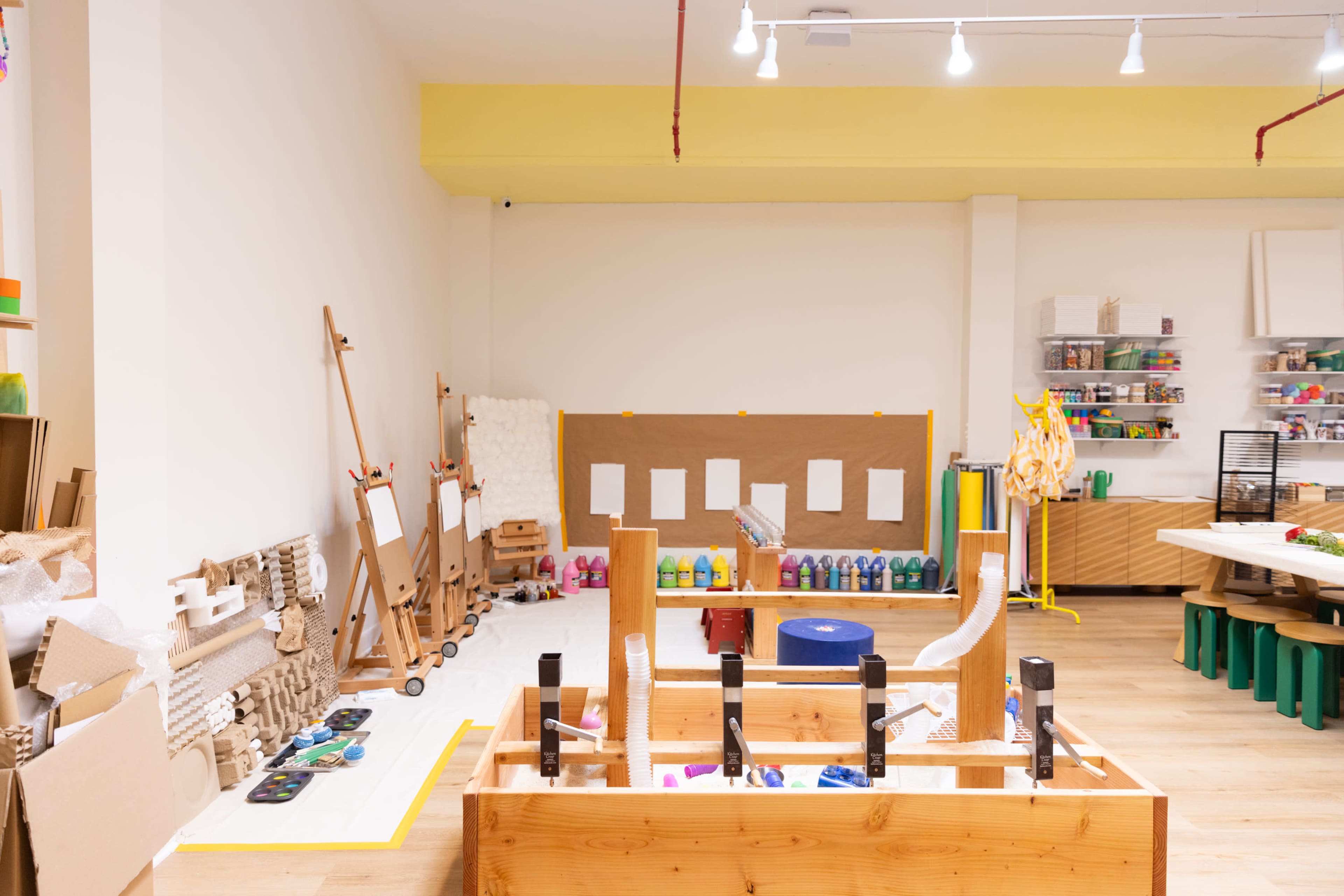 The image shows a brightly lit art and craft room featuring wooden tables, easels, and various art supplies organized on shelves.