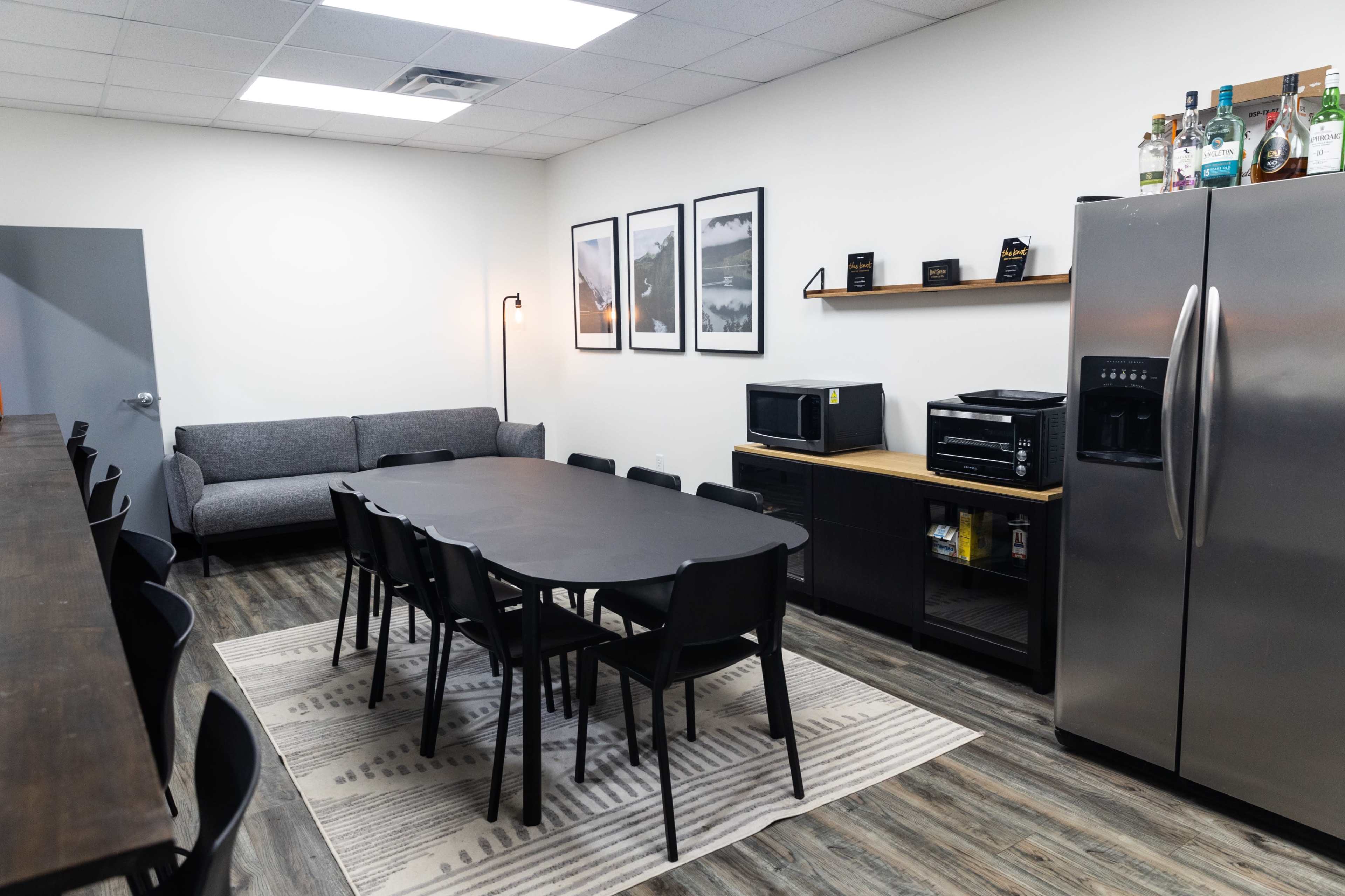 The image shows a modern break room with a long black table surrounded by chairs, a couch, kitchen appliances, and framed artwork on the walls.