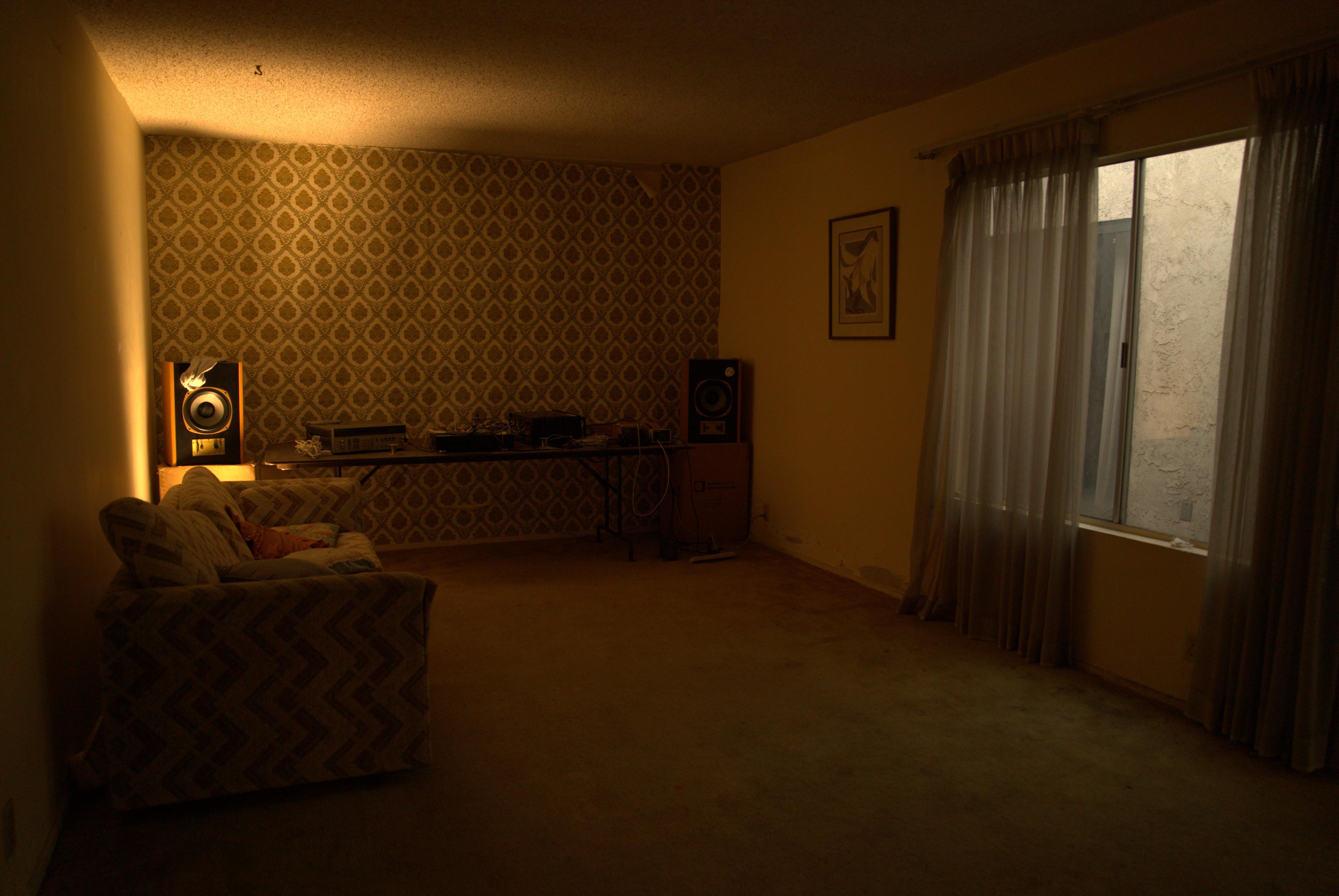 A sparse living room features a patterned wallpaper, a couch, a table with speakers, and a window with sheer curtains.