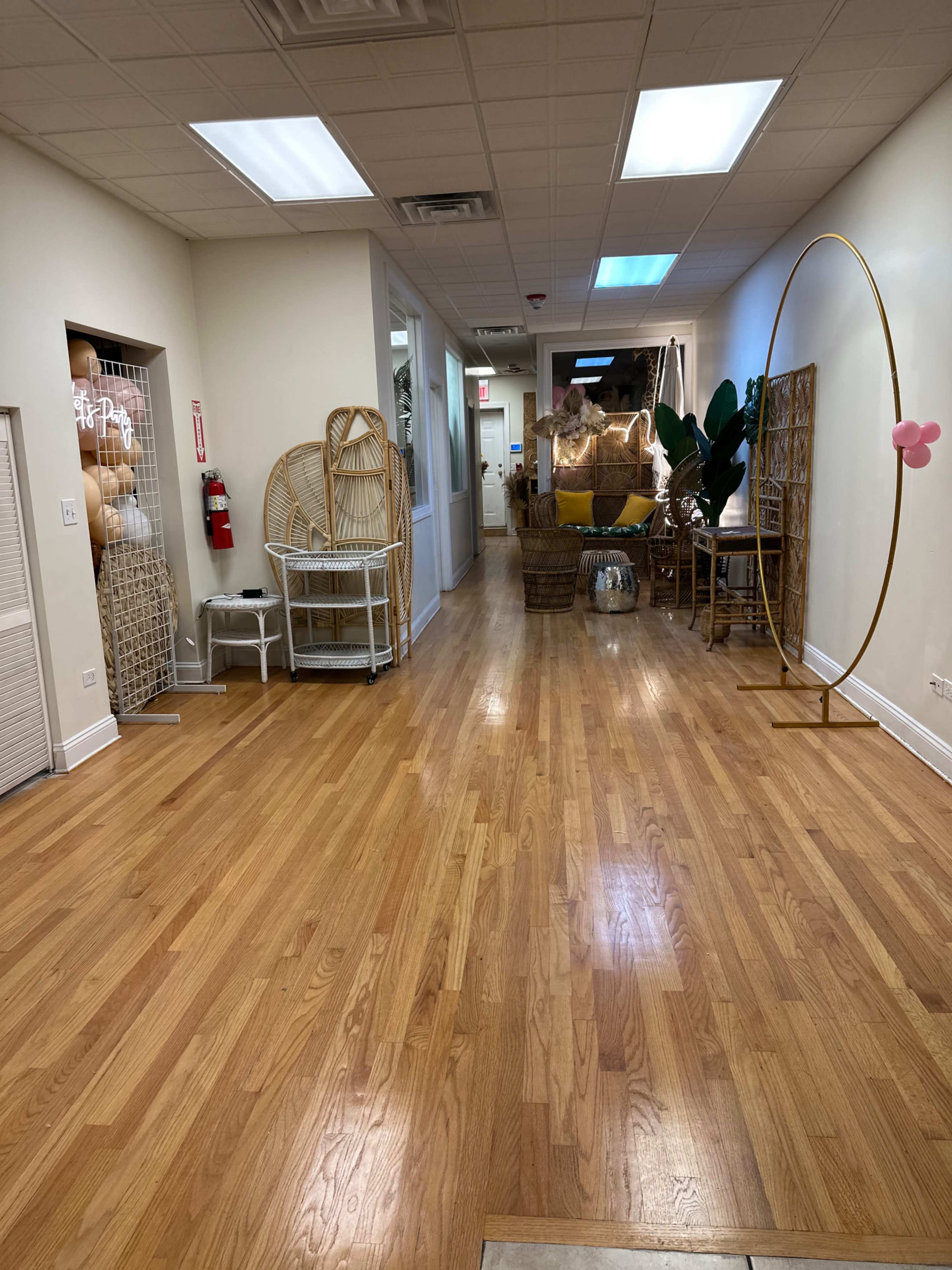 The image shows a long hallway with polished wooden floors, flanked by various decorative items, including plants and wicker furniture, leading to a brightly lit area at the far end.