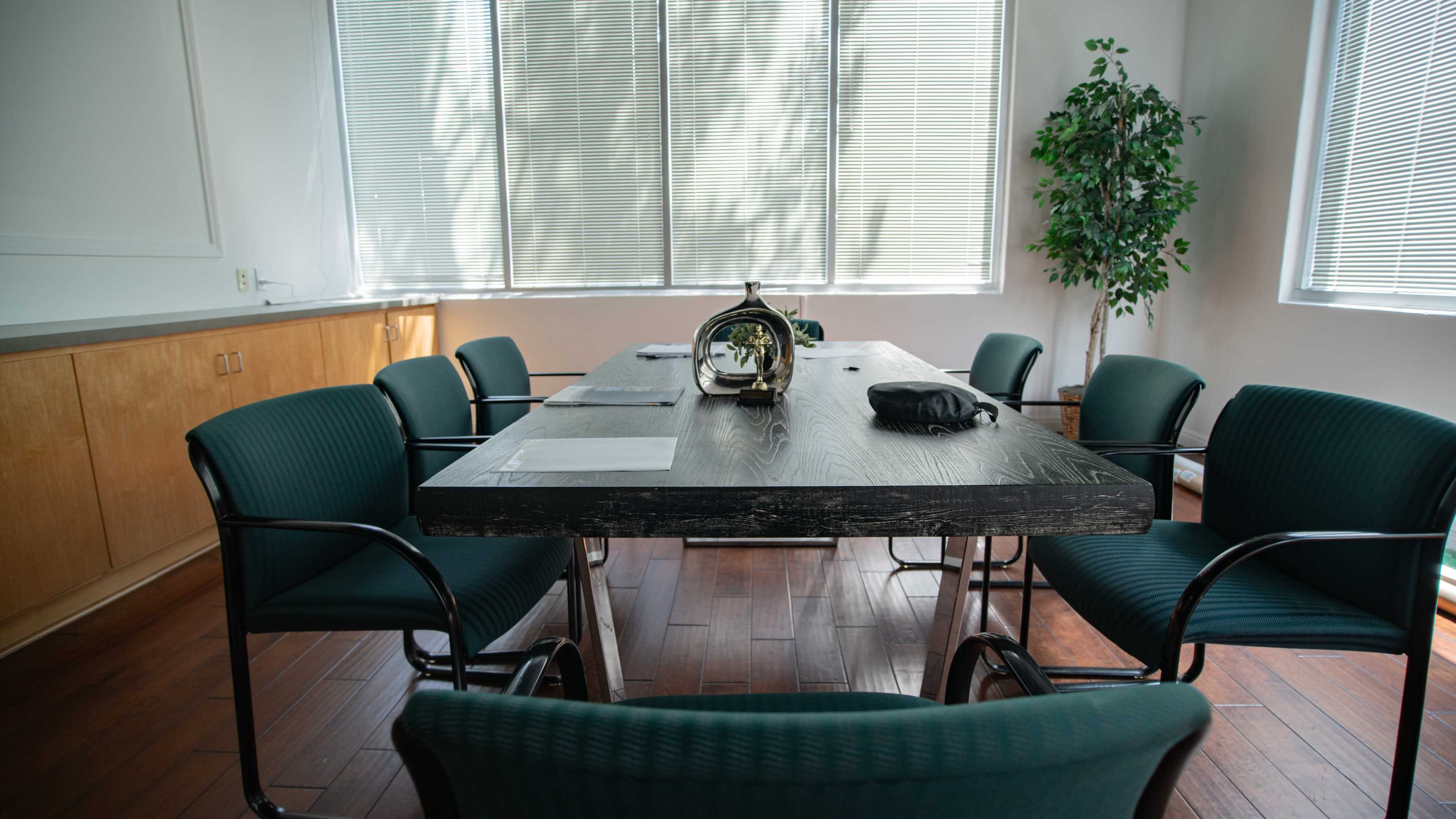A conference room features a large wooden table surrounded by six green chairs and a plant in the corner, with natural light filtering through blinds.