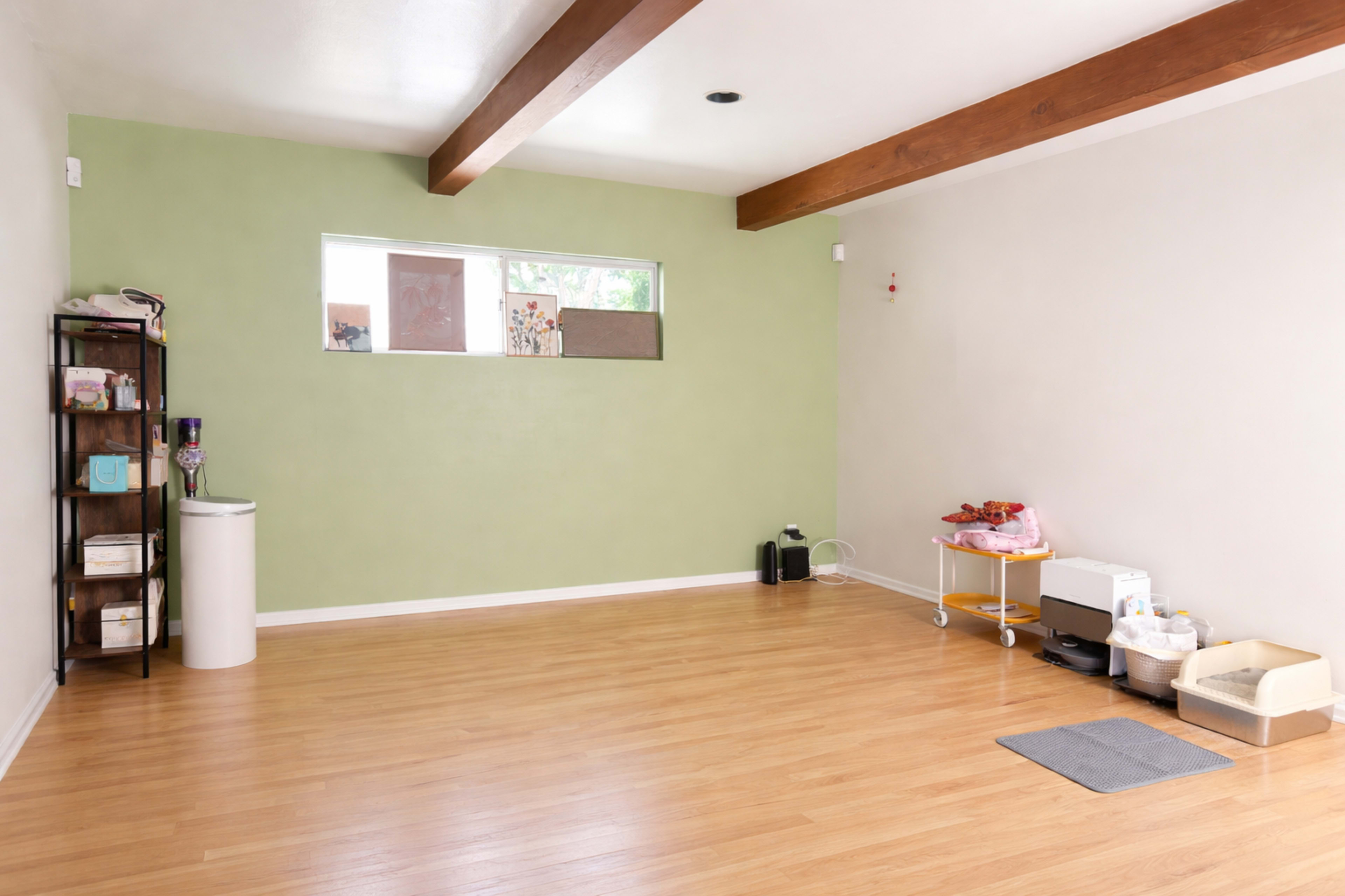 The image shows a spacious, well-lit room with light wood flooring, a green accent wall, and minimal furnishings including a shelf and various objects in the corners.