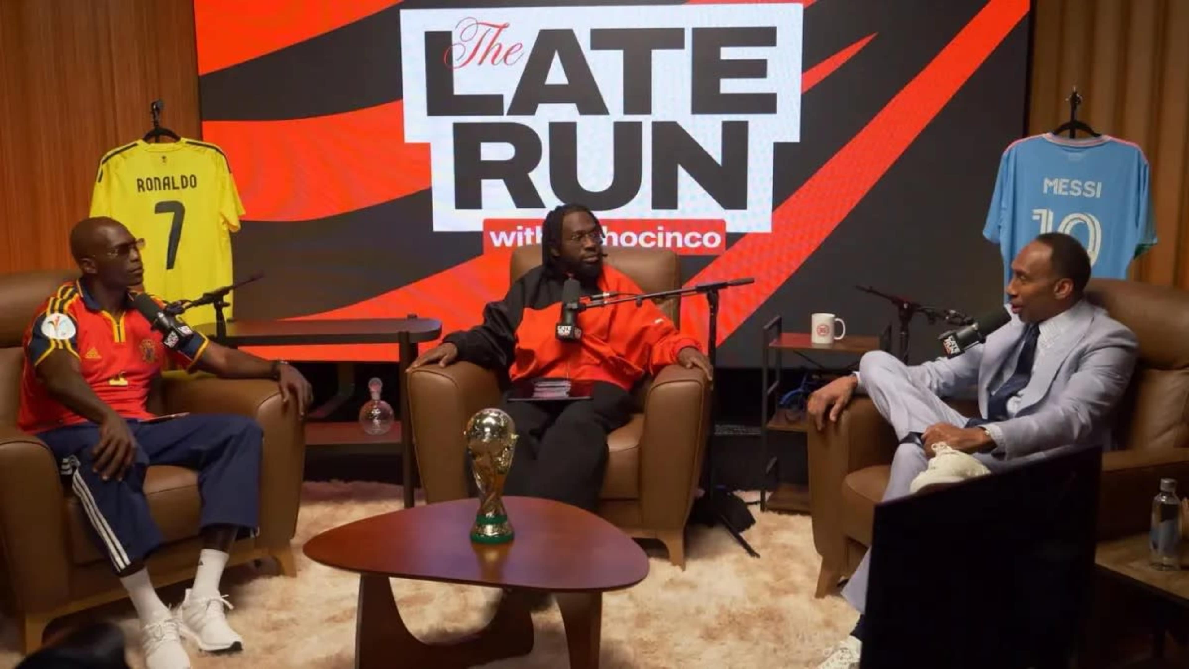 Three men sit in armchairs in a studio decorated with sports jerseys and a soccer trophy, discussing topics related to sports and media.