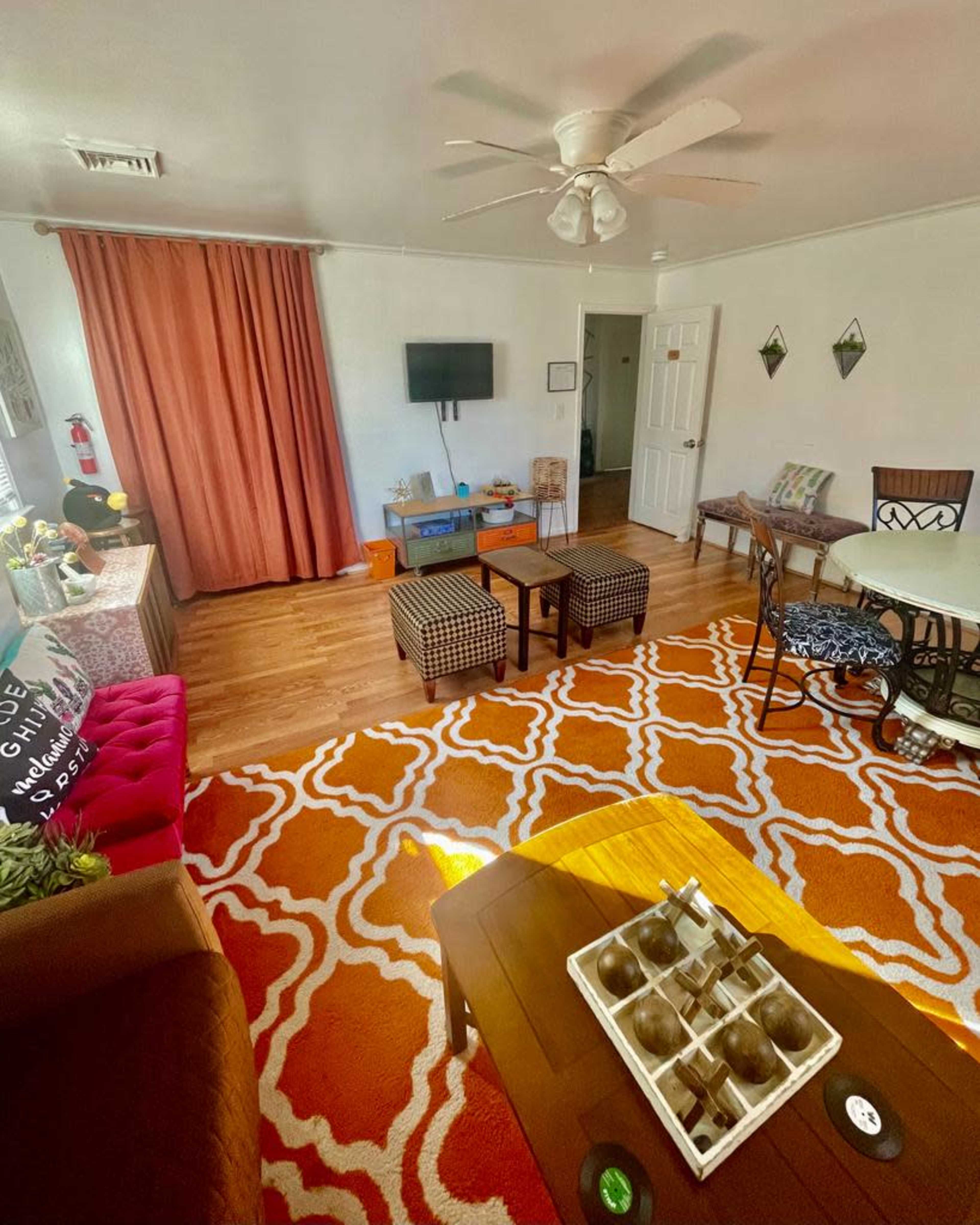 The room features a patterned orange and white rug, a pink couch, a round dining table, and a flat-screen TV on the wall.